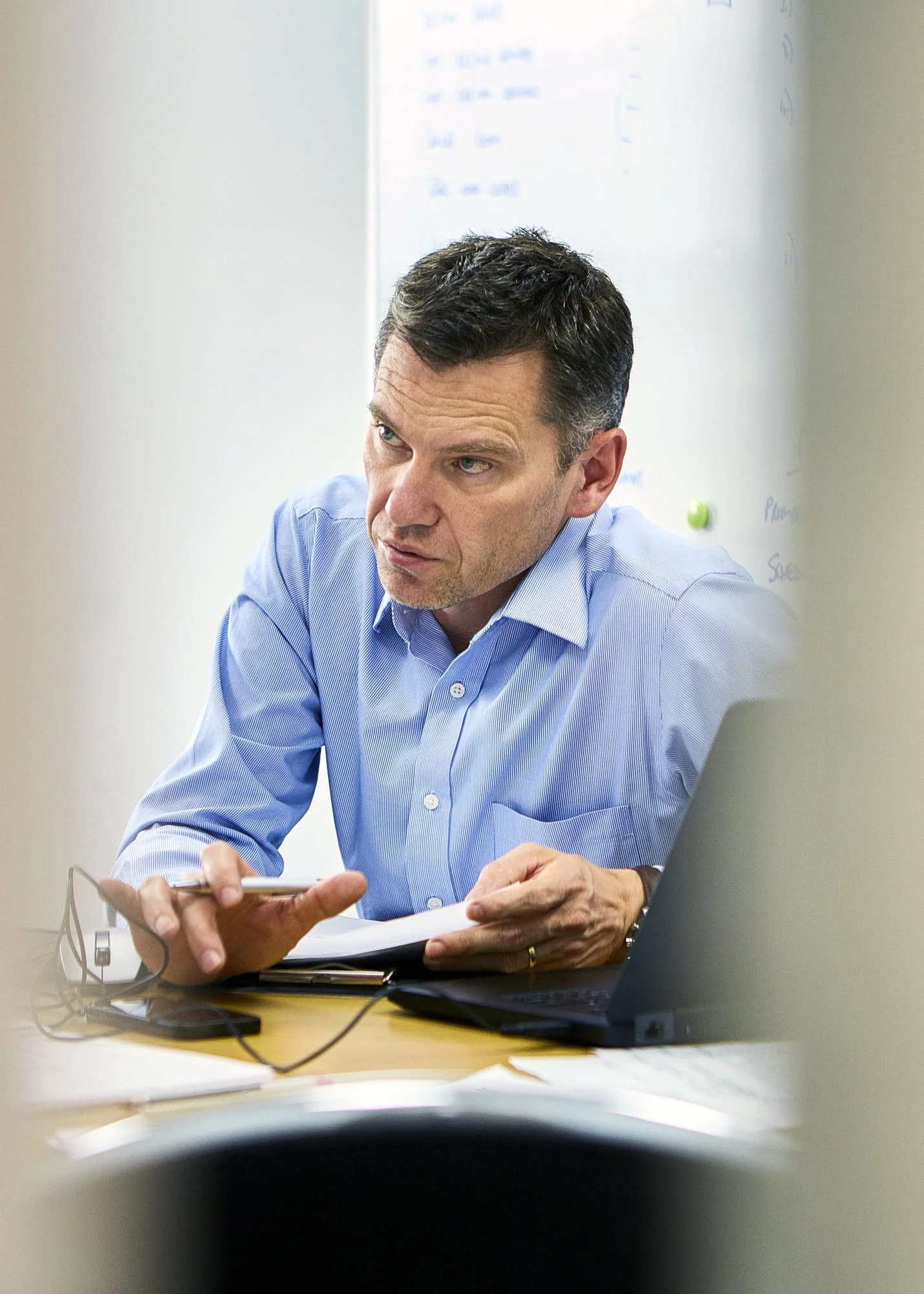 A man in a blue dress shirt sitting at a desk with various electronic devices and papers, looking to his right and holding a smartphone.