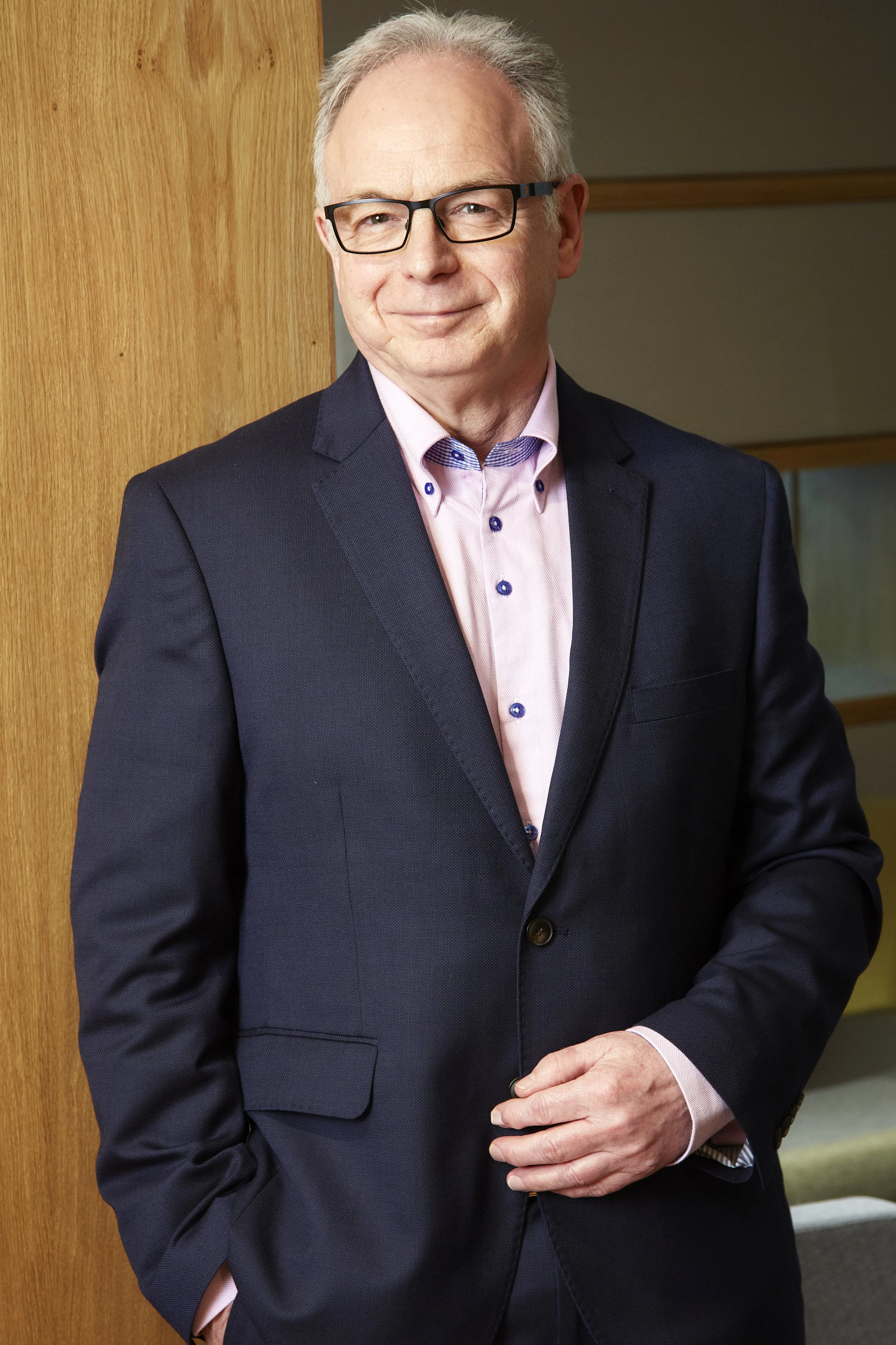 A middle-aged man with glasses and gray hair wearing a dark suit and light pink shirt, standing indoors near a wooden wall, smiling slightly.