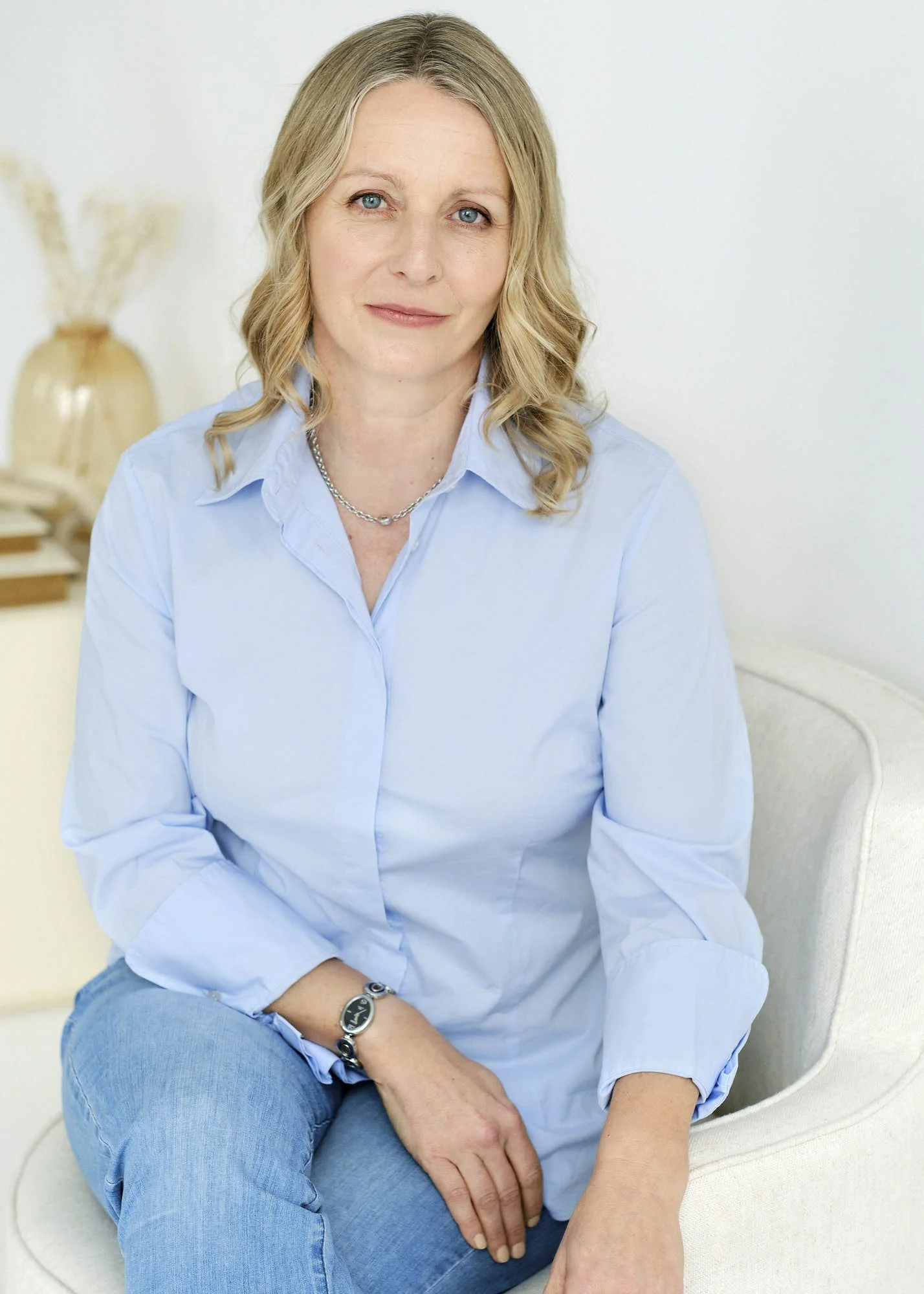 A woman with blonde wavy hair, blue eyes, wearing a light blue button-up shirt, jeans, a silver chain necklace, a black watch, and a bracelet, sitting on a light-colored armchair with her hand resting on her lap, in a bright room with minimal decor.