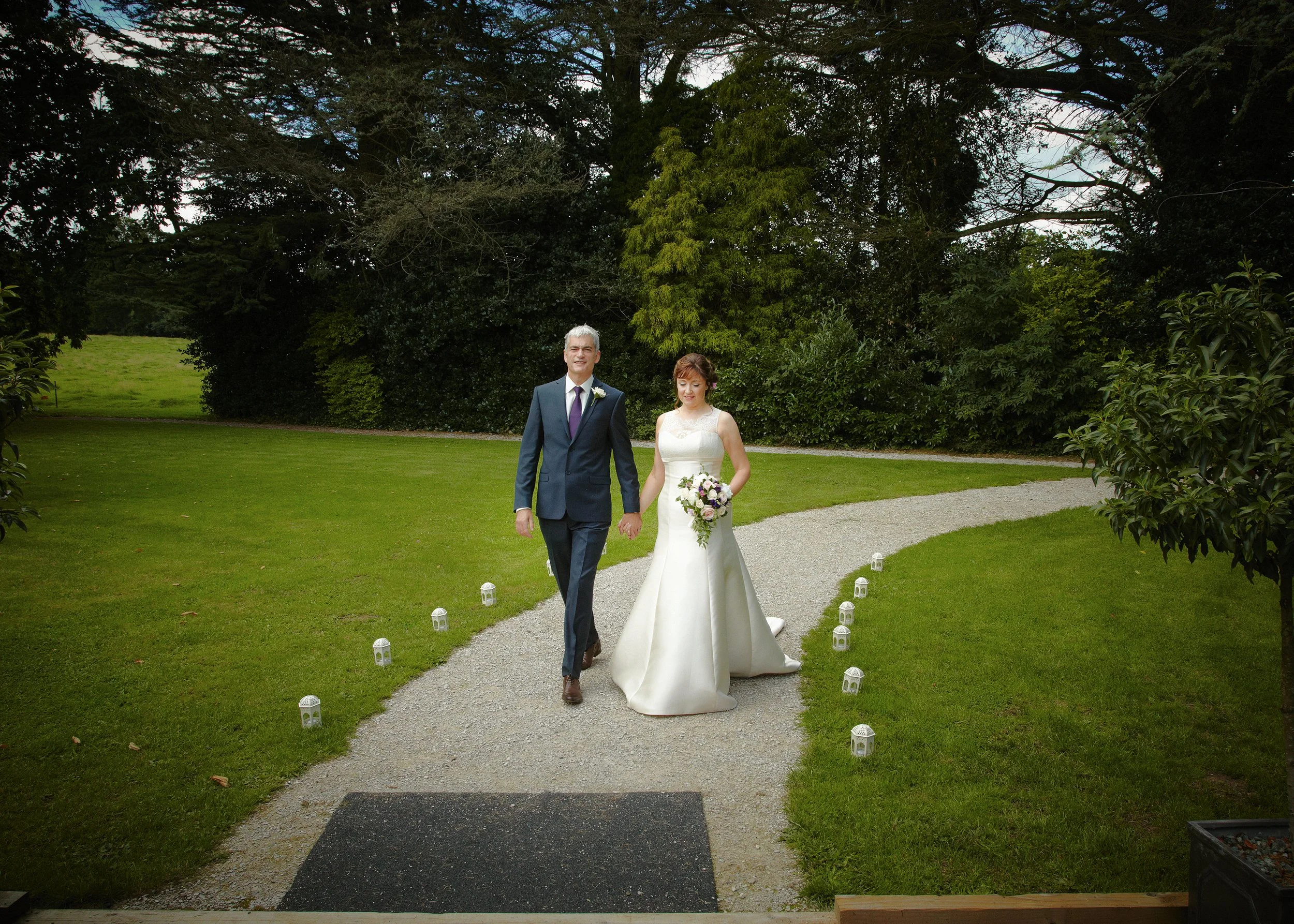 A bride and groom walking hand in hand on a gravel path at an outdoor wedding ceremony, surrounded by green trees and grass, with small lanterns along the path.