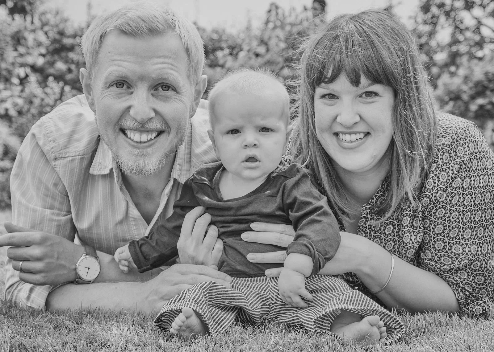 Black and white photograph of a smiling, happy family of three lying on the grass outdoors. The father, with light hair and a beard, is on the left, the mother, with dark hair, is on the right, and their young child is in the middle, looking curious with a slightly confused expression.