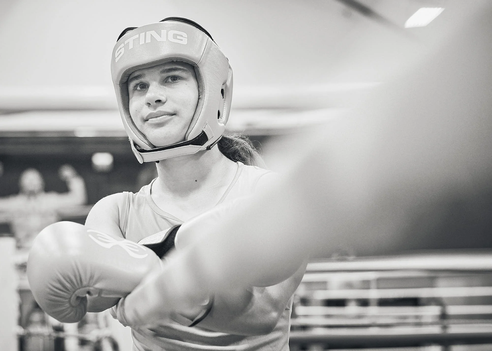 A young woman wearing boxing gear, including a head guard and gloves, practicing or sparring in a gym.
