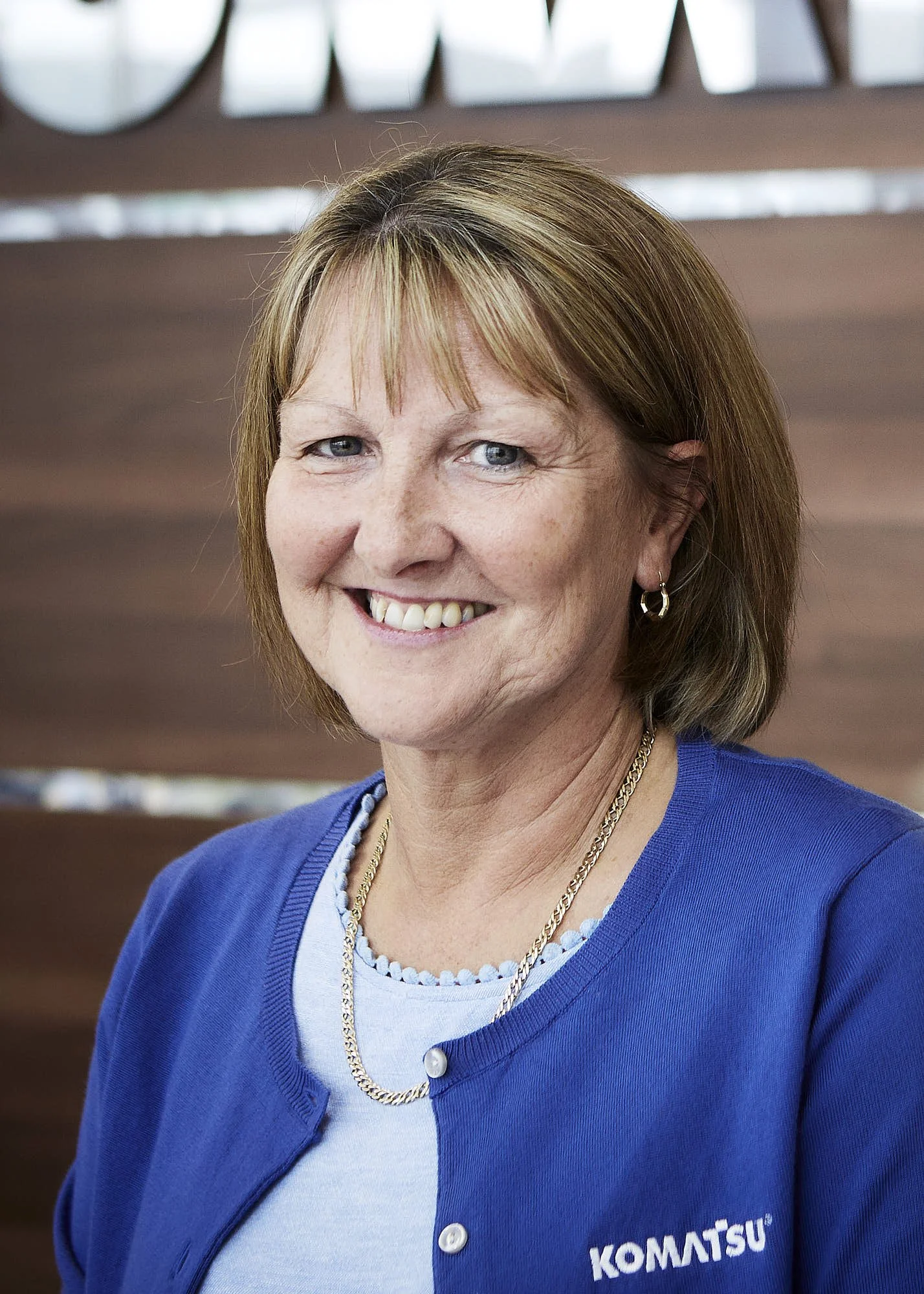 A middle-aged woman with short light brown hair wearing a blue jacket and a white shirt, smiling in front of a wooden background.