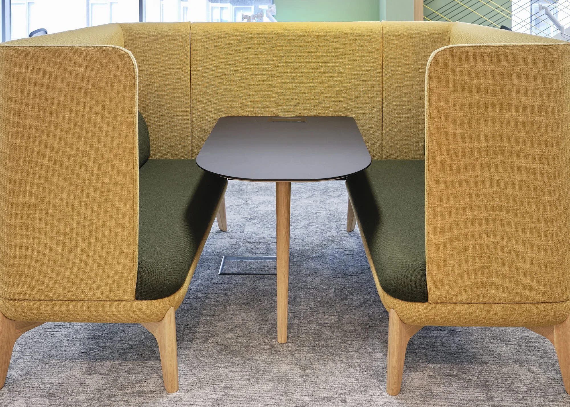 Two mustard-yellow booths with curved armrests and black cushions, separated by a small black table with wooden legs, located on a gray carpet in an open, modern office space with large windows and green accents.