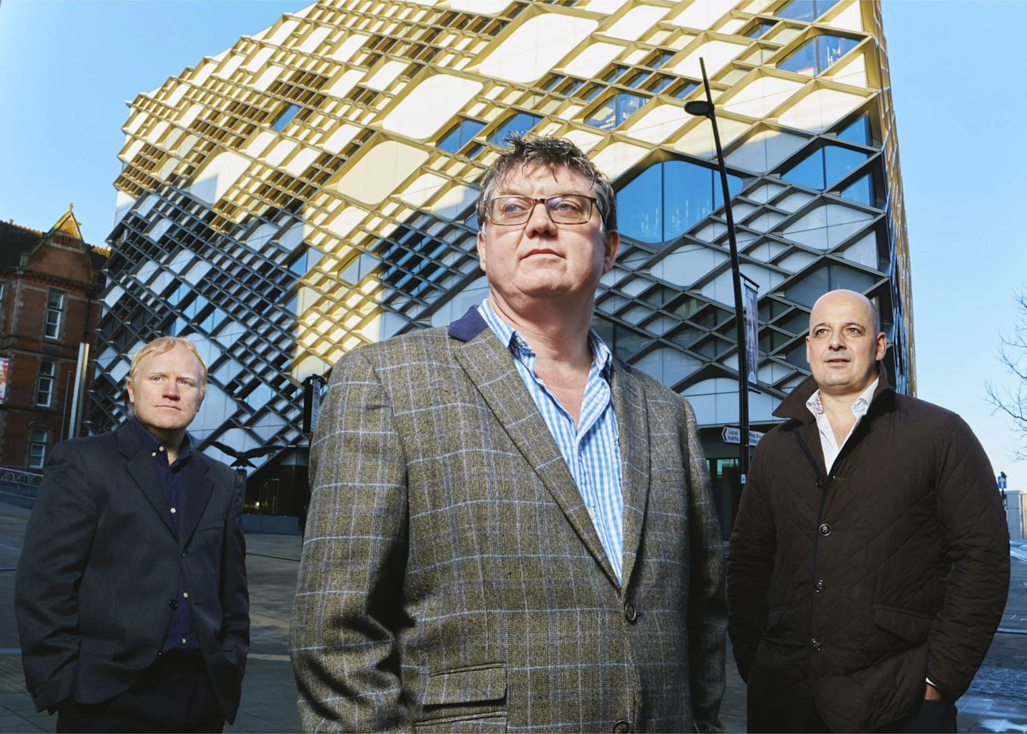 Four men standing outdoors in front of a modern glass and gold building, with a blue sky above.