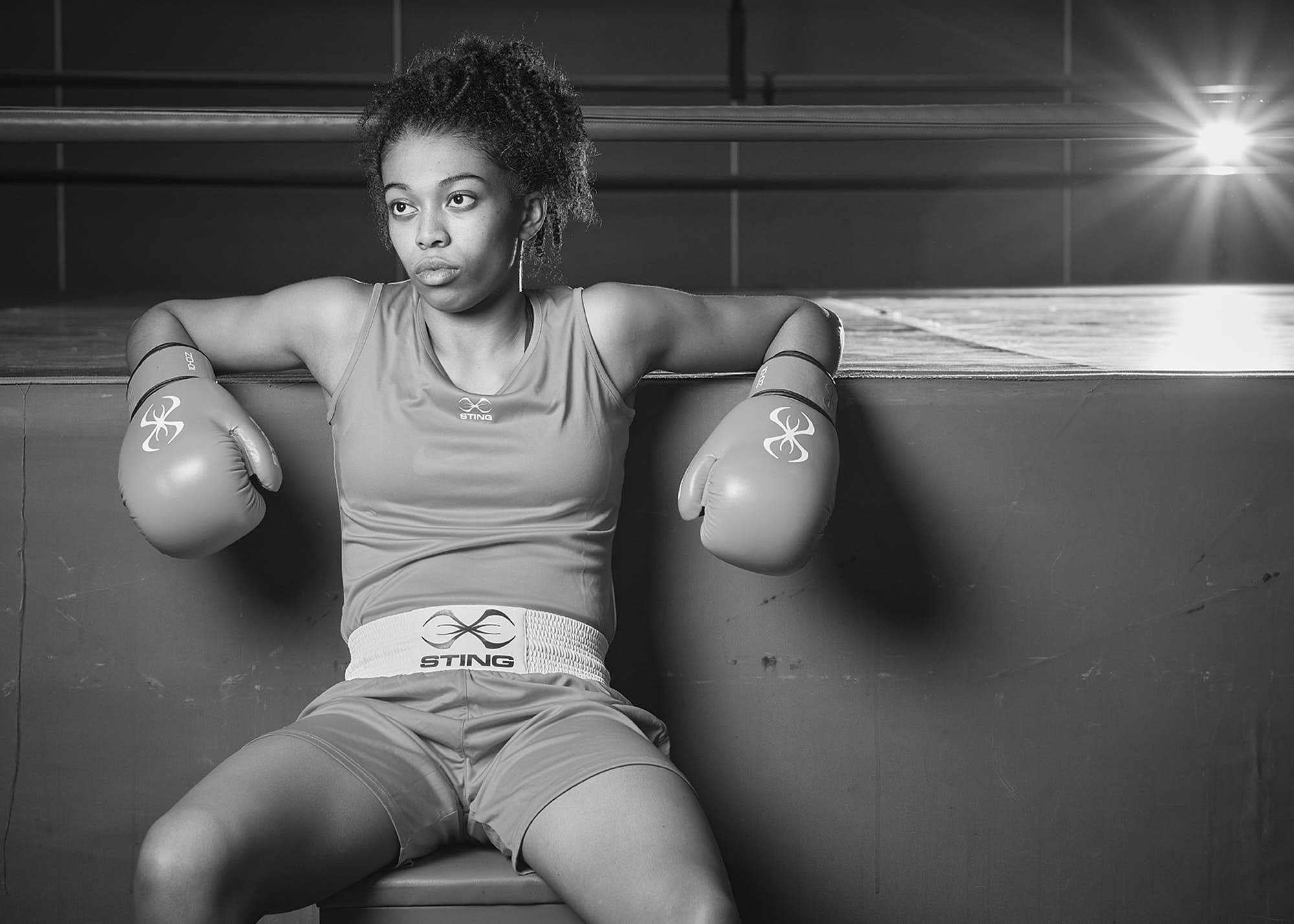 Black and white photo of a female boxer sitting against a gym wall with boxing gloves on her hands, looking off to the side with a determined expression.