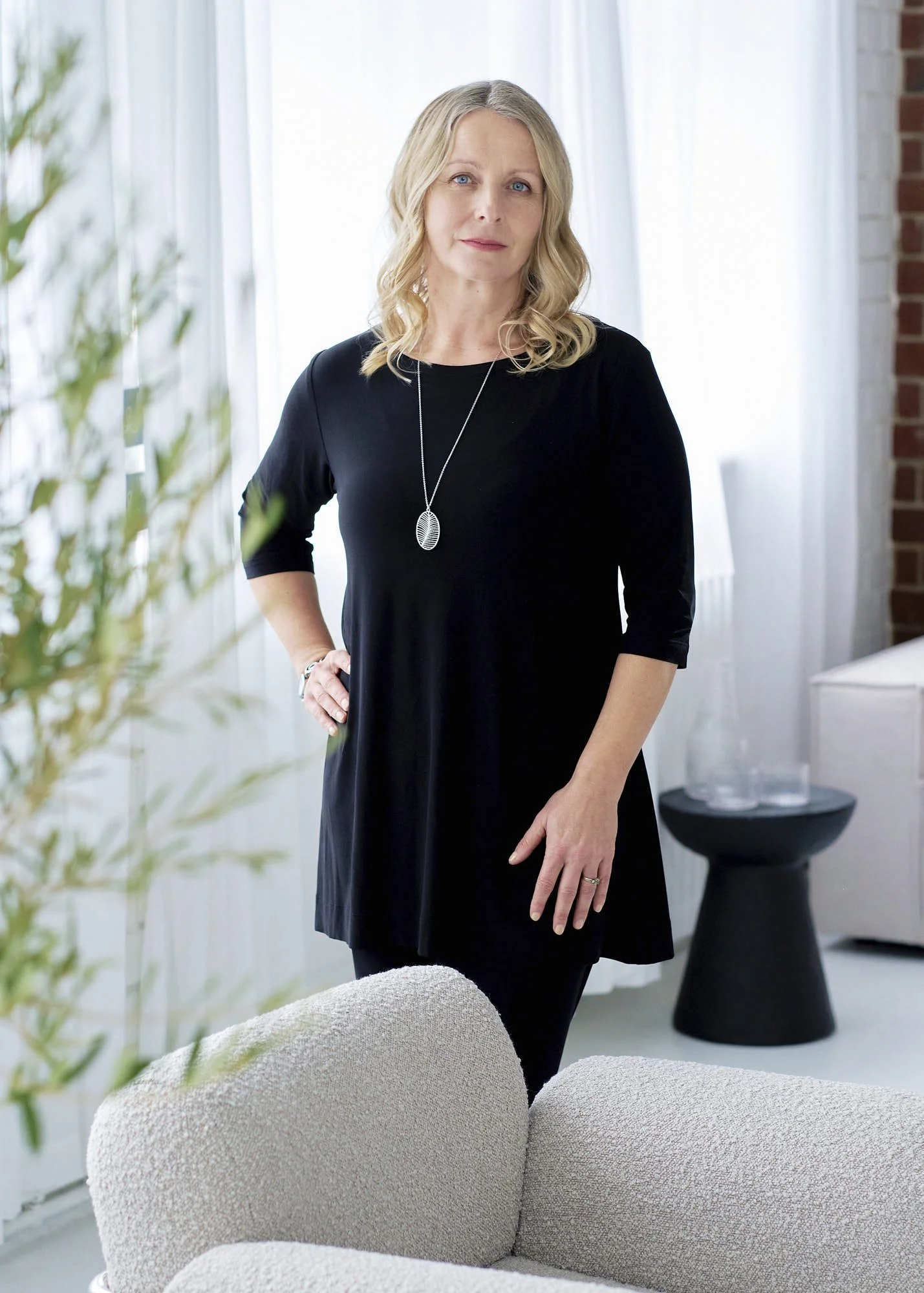 A middle-aged woman with blond hair, wearing a black dress and a necklace, standing in a bright, modern living room with white curtains and a brick wall in the background.