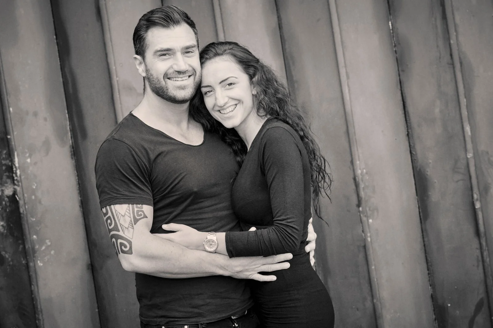A smiling couple embracing each other against a wooden wall, in black and white.