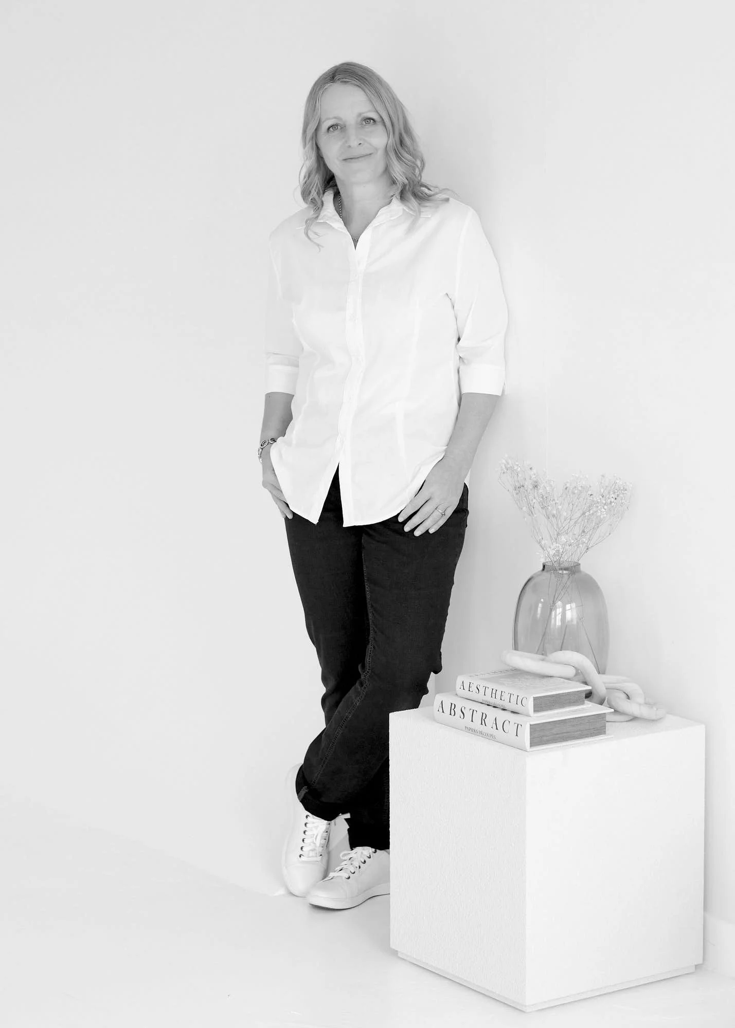 Black and white photo of a woman with wavy hair, wearing a white button-up shirt, dark jeans, and white sneakers, standing with her hands in pockets next to a white pedestal with books and a vase with dried flowers on top.