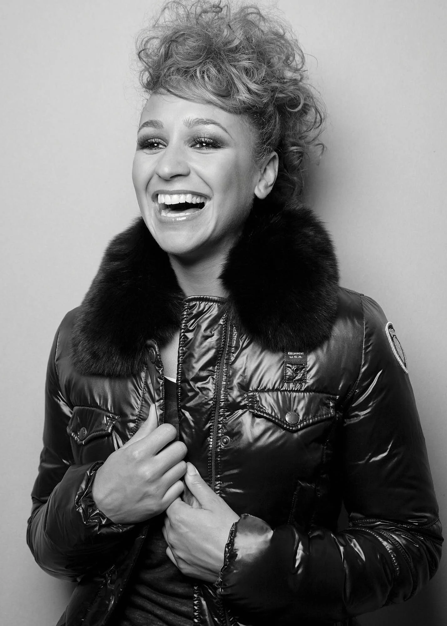 Black and white photo of a woman smiling, wearing a shiny puffer jacket with fur collar, with her hand grasping another person's hand.