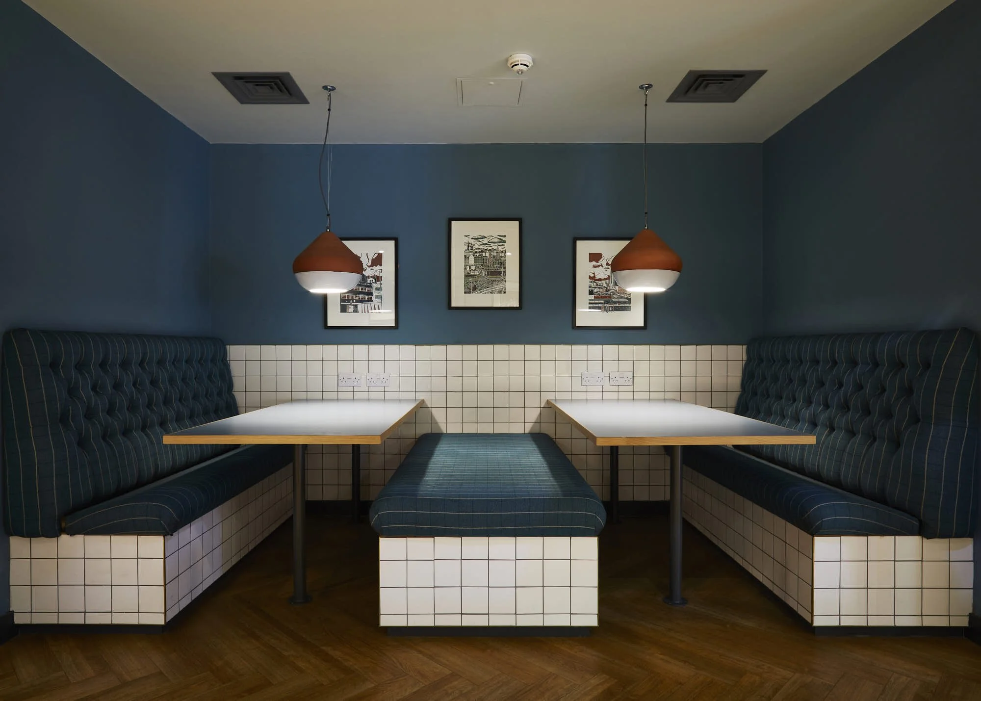 Booth seating area in a restaurant with blue padded benches, white tiled bases, and white tabletops. Three framed black-and-white pictures hang on a blue wall. Two hanging lamps with brown shades illuminate the space.