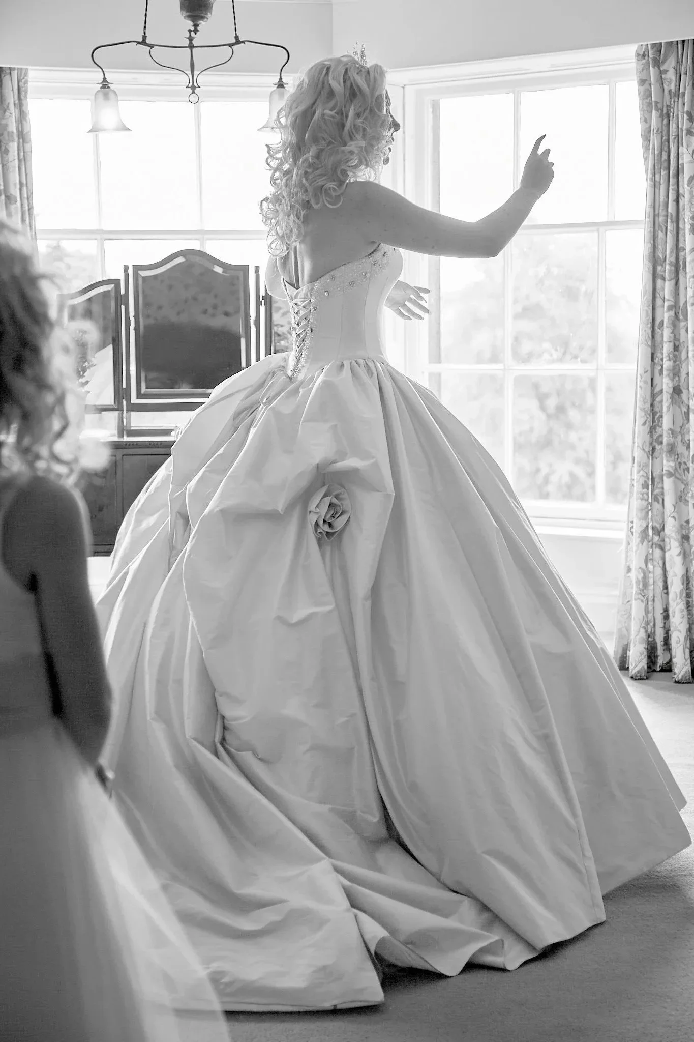 A woman in a wedding dress is standing in front of a large window, with her right arm raised and her finger pointing upwards. The dress has a full skirt with a large flower decoration, and she has long, curly hair. The room has patterned curtains and a chandelier hanging from the ceiling.
