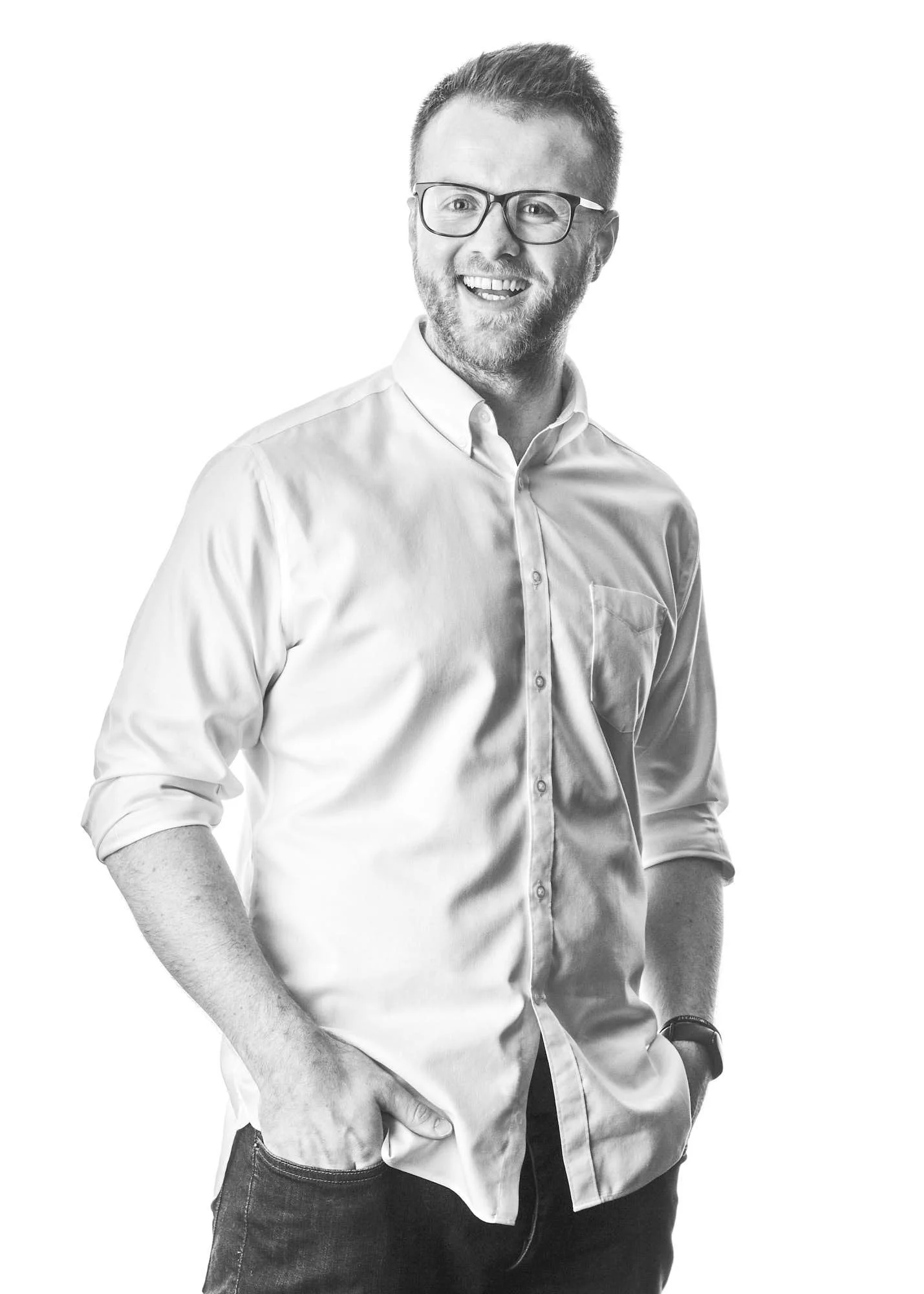 Black and white photo of a man wearing glasses, a button-up shirt, and a wristwatch, smiling and posing with hands in pockets against a plain background.