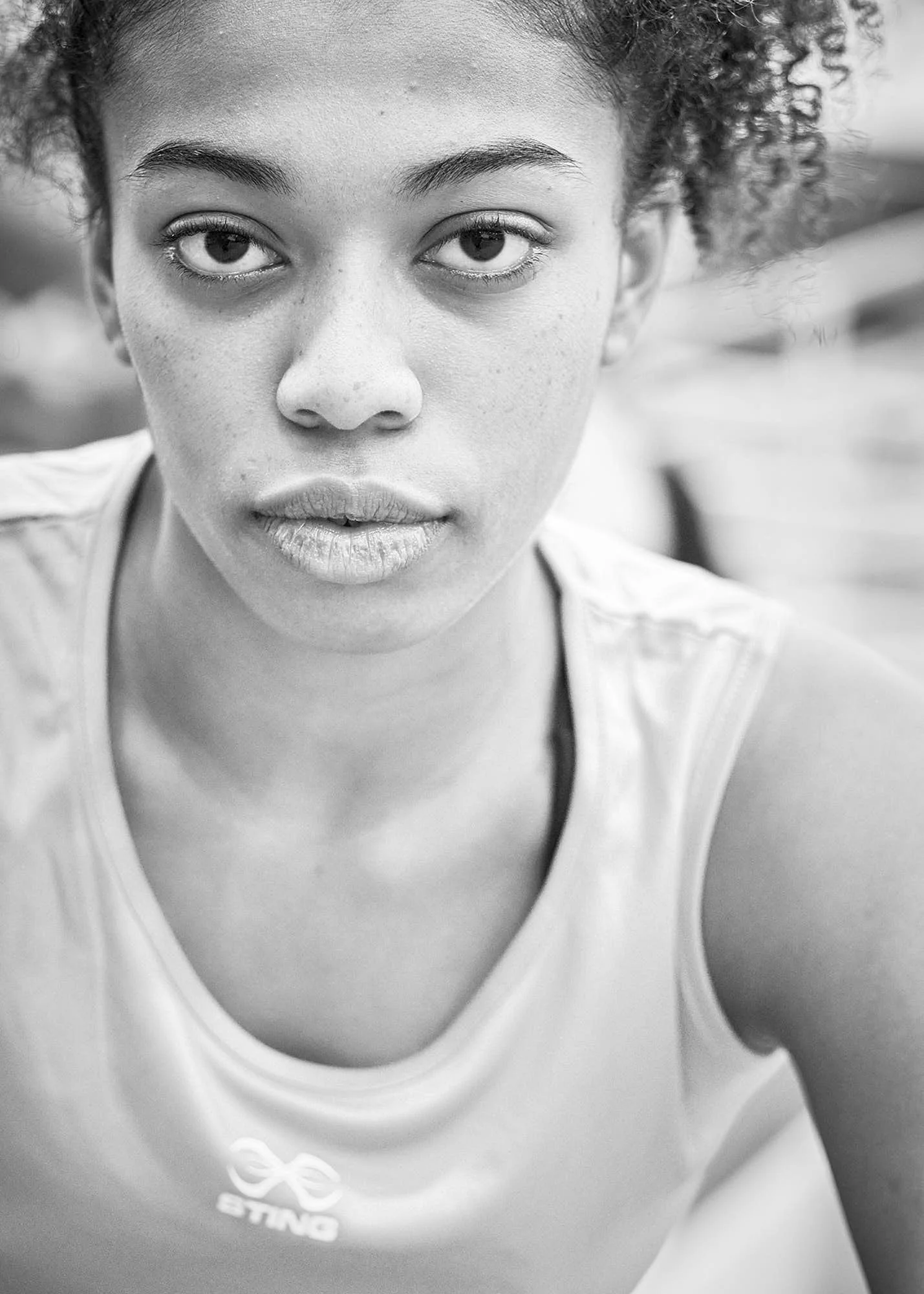 Close-up black and white photo of a young woman with curly hair, wearing a white tank top with a logo, looking directly into the camera with a neutral expression.