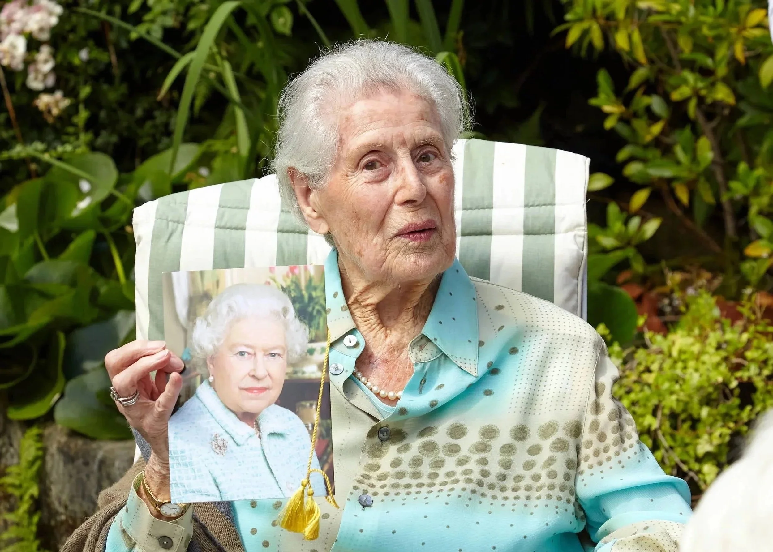 Elderly woman with white hair sitting outdoors, holding a photo of herself when she was younger, surrounded by green foliage.