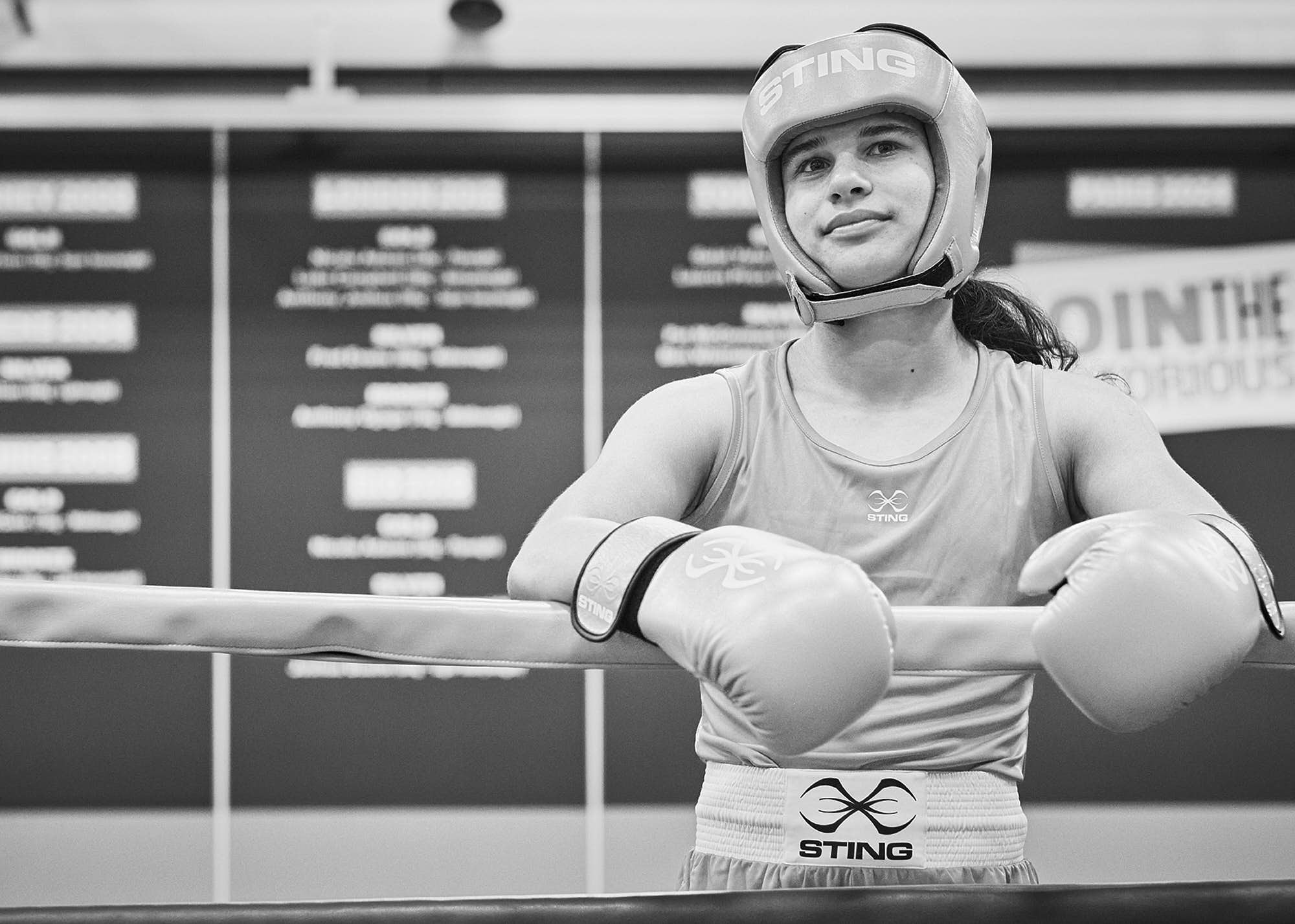 Black and white photo of a female boxer in a boxing gym, wearing a helmet and boxing gloves, resting with her arms crossed on the boxing ring ropes.