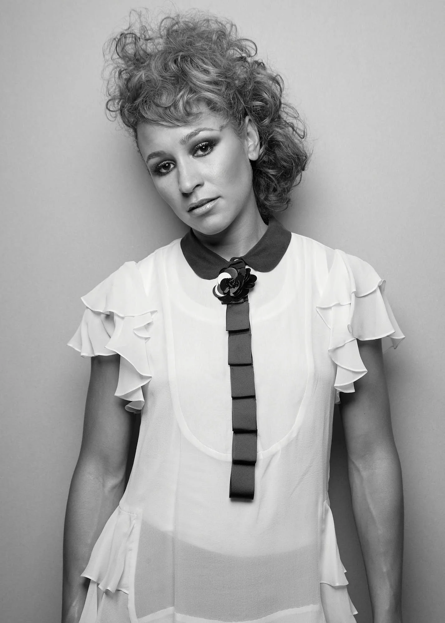 Black and white photo of a woman with curly hair, wearing a blouse with ruffled sleeves and a decorative flower on the collar, standing against a plain background.