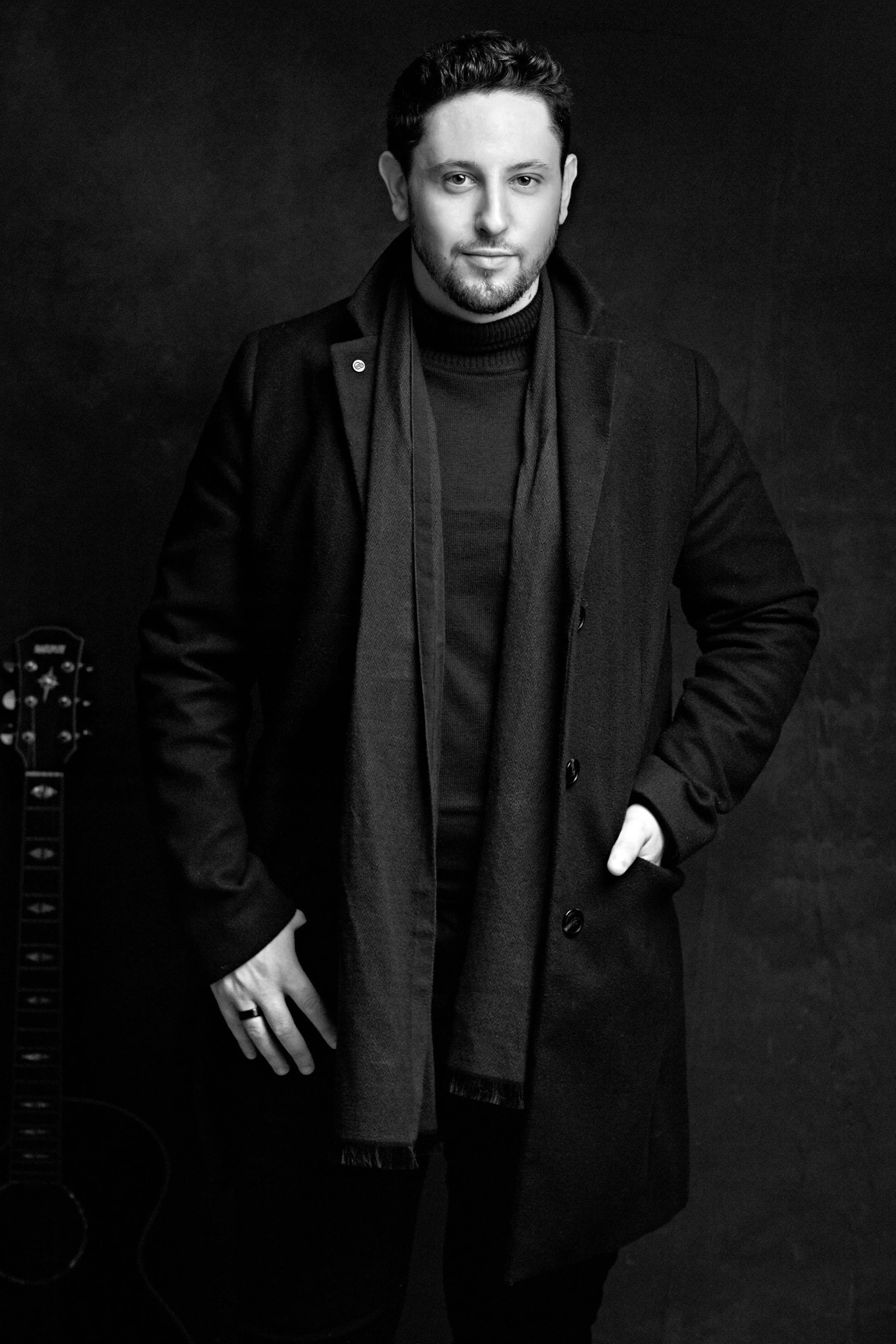 Black and white portrait of a man with short dark hair and a beard, wearing a dark overcoat and a black turtleneck, standing against a dark background.