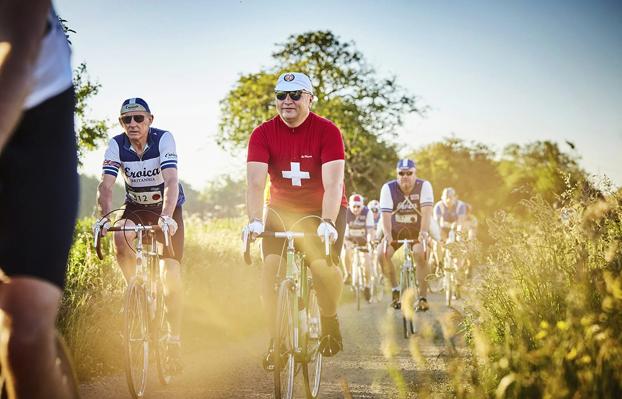 A group of cyclists riding on a dirt trail outdoors during sunset, with trees and greenery on either side, wearing colorful jerseys and sunglasses.