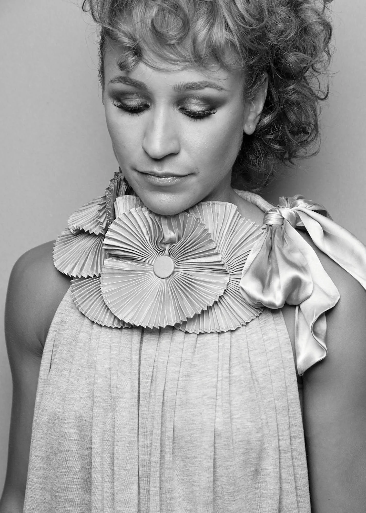 A woman with curly hair looking downward, wearing a decorated top with ruffled details and a satin ribbon tied at the shoulder.
