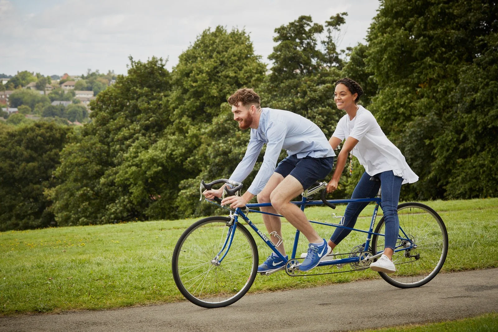 A young couple riding a tandem bicycle on a grassy park path, with trees and distant houses in the background.