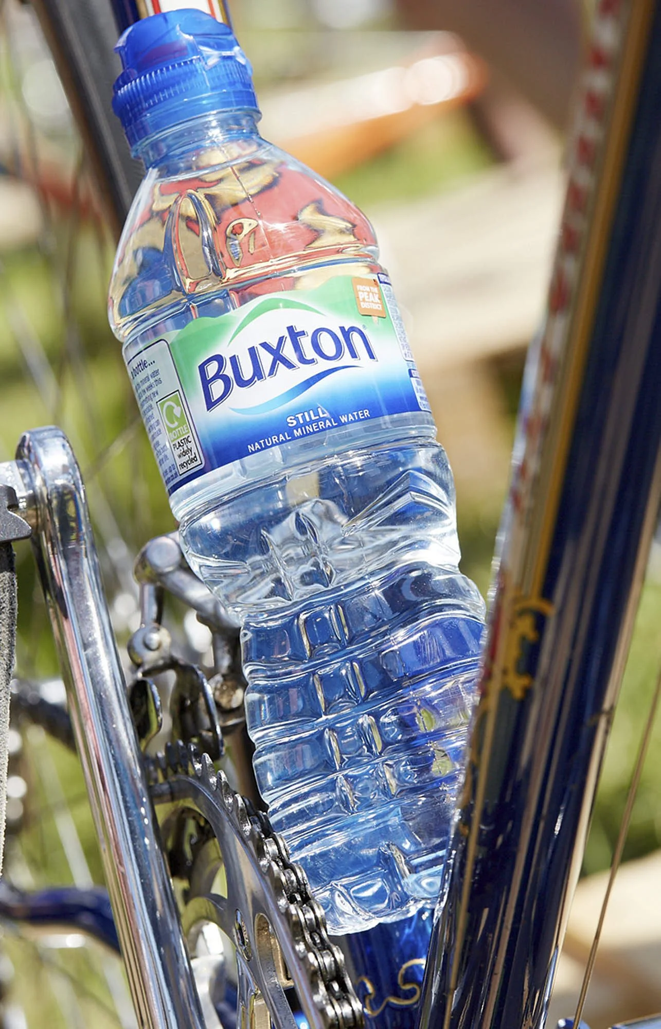 A plastic water bottle labeled 'Buxton Still Natural Mineral Water' placed in a bicycle bottle holder on a bike with a chain and gears visible.