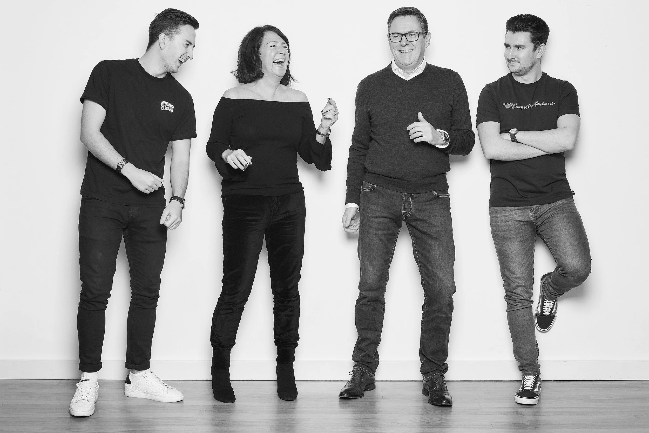 A group of five people standing against a plain wall, laughing and talking, dressed casually in dark clothing, with one woman in the middle wearing an off-the-shoulder top.