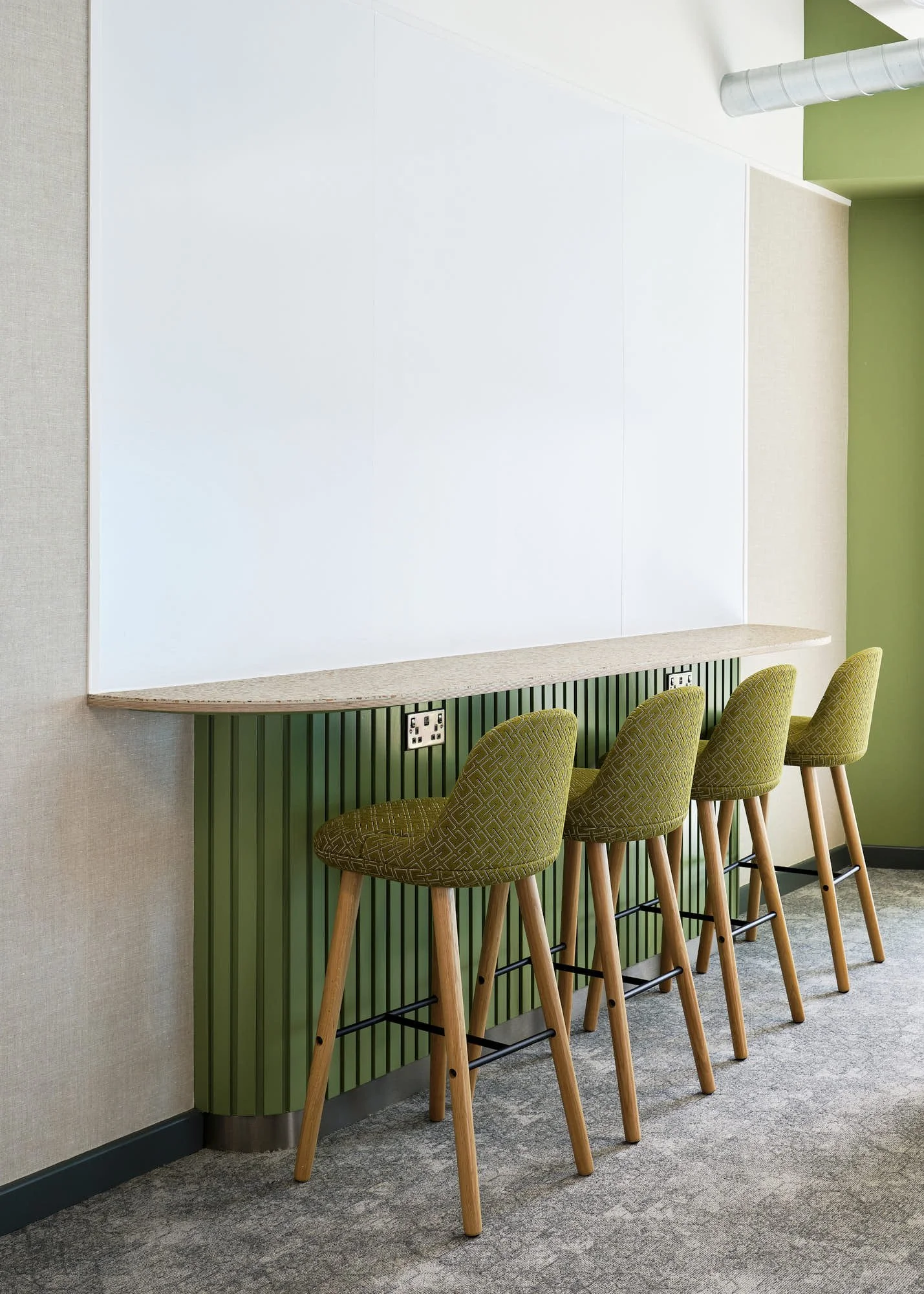 A row of three green upholstered bar stools with wooden legs, positioned against a green and beige wall with a whiteboard above. There are electrical outlets beneath the whiteboard.