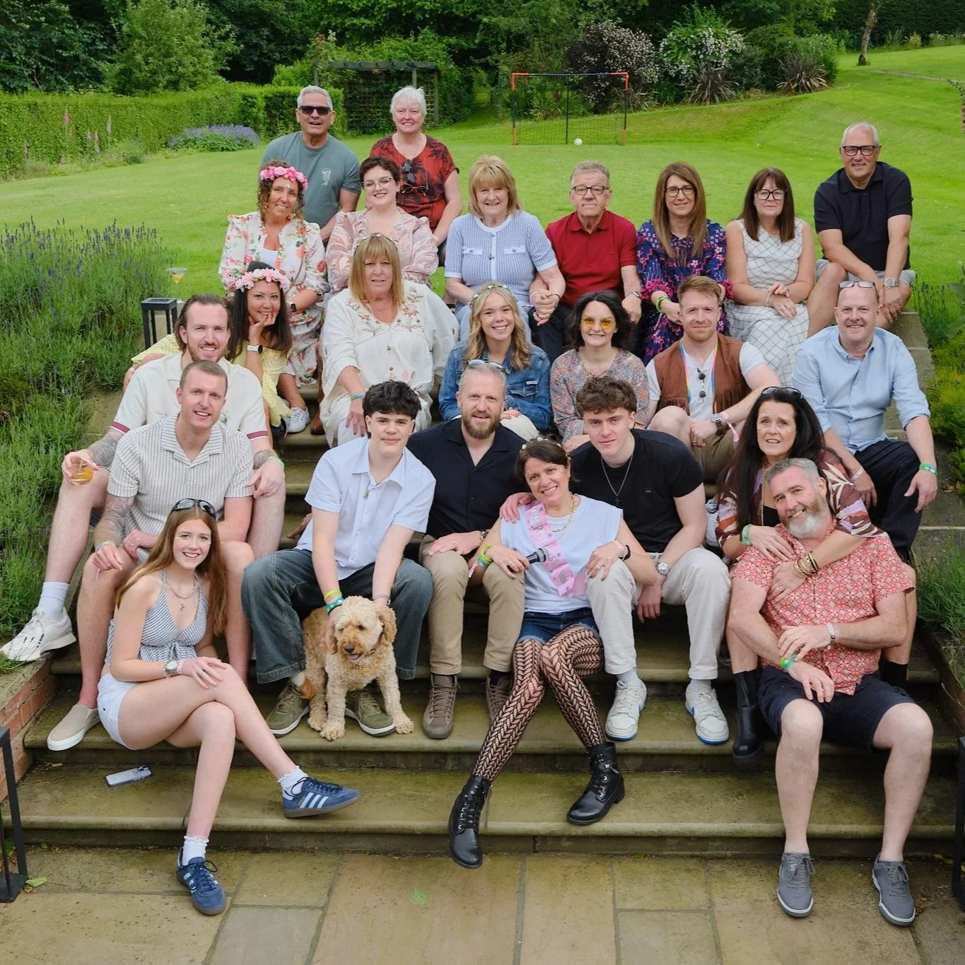 A large group of people sitting on outdoor steps in a garden with green grass and trees, with a soccer goal and a white ball in the background. There is a dog in front of the group.