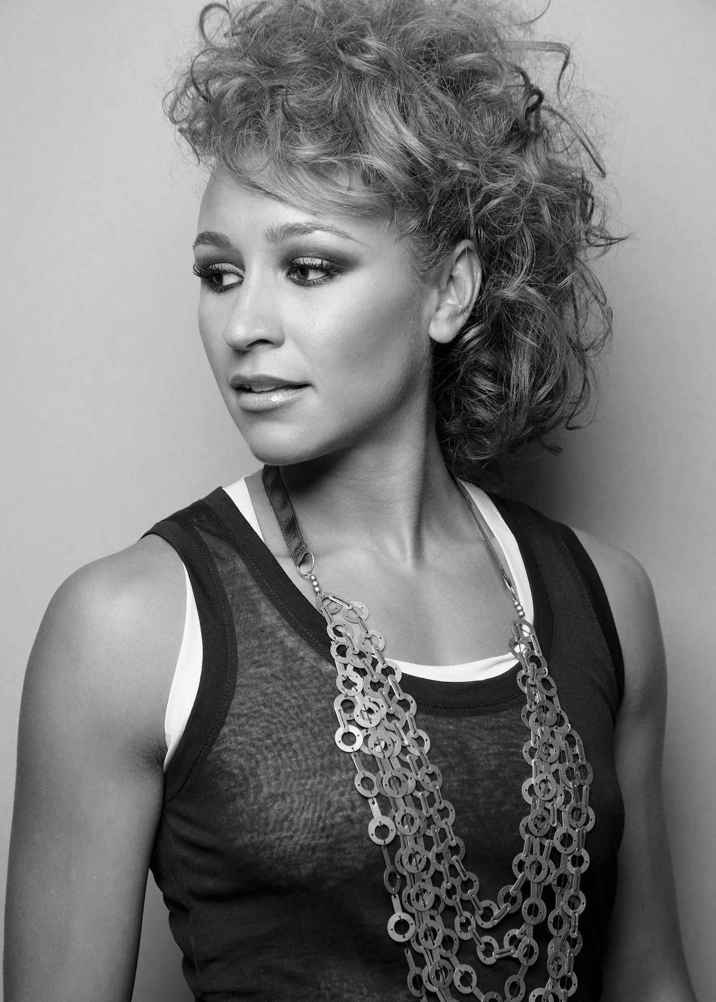 Black-and-white portrait of a woman with curly hair styled in an updo, wearing a sleeveless top and layered chain necklaces.