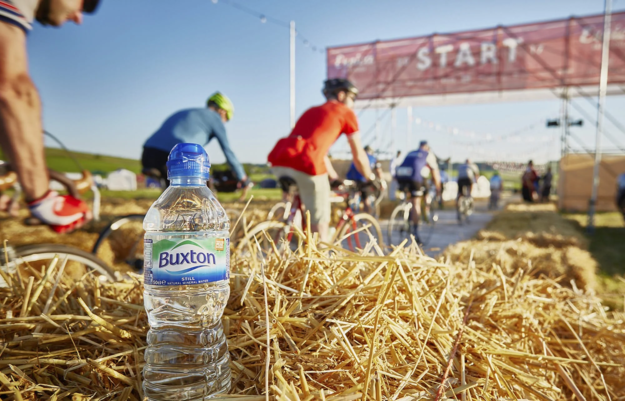 A plastic bottle of Buxton still water placed on a bed of straw at the starting line of a bike race event, with cyclists and a large red start banner in the background.