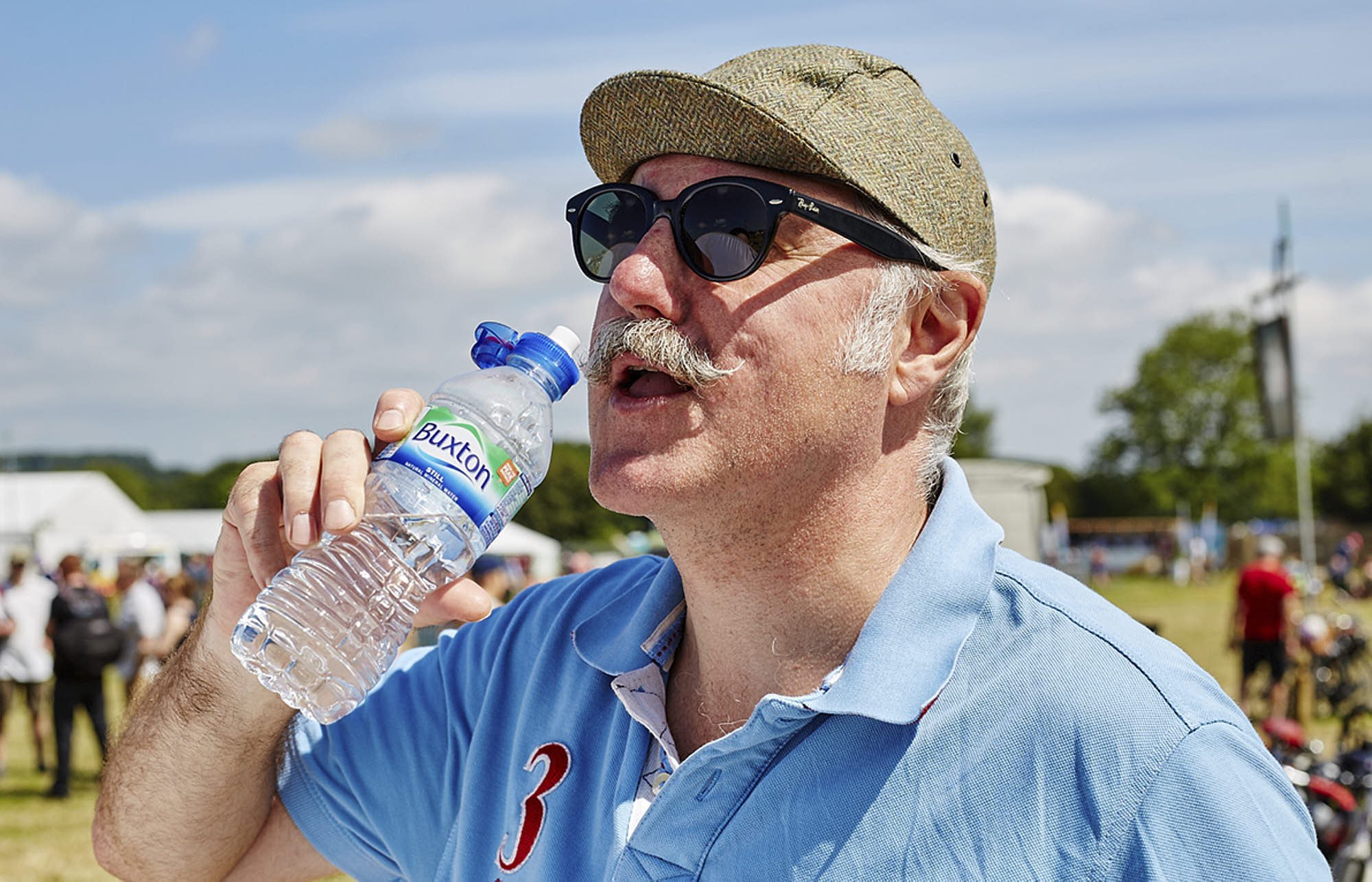 A man wearing sunglasses, a beige cap, and a light blue polo shirt is drinking water from a plastic bottle at an outdoor event, with a crowd and cloudy sky in the background.