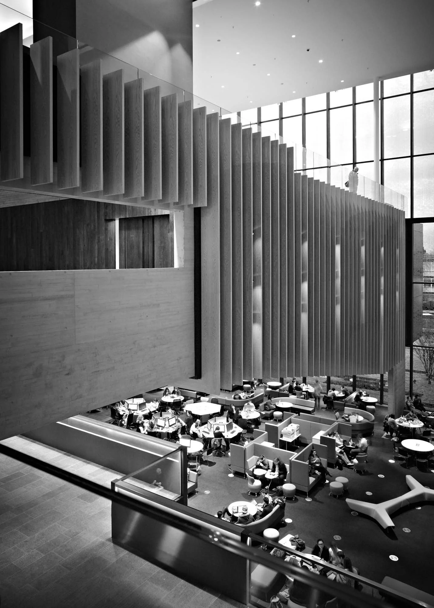 Interior view of a modern library or study area with people working at tables and a large staircase with wooden slats leading to an upper level, having high glass windows letting in natural light.