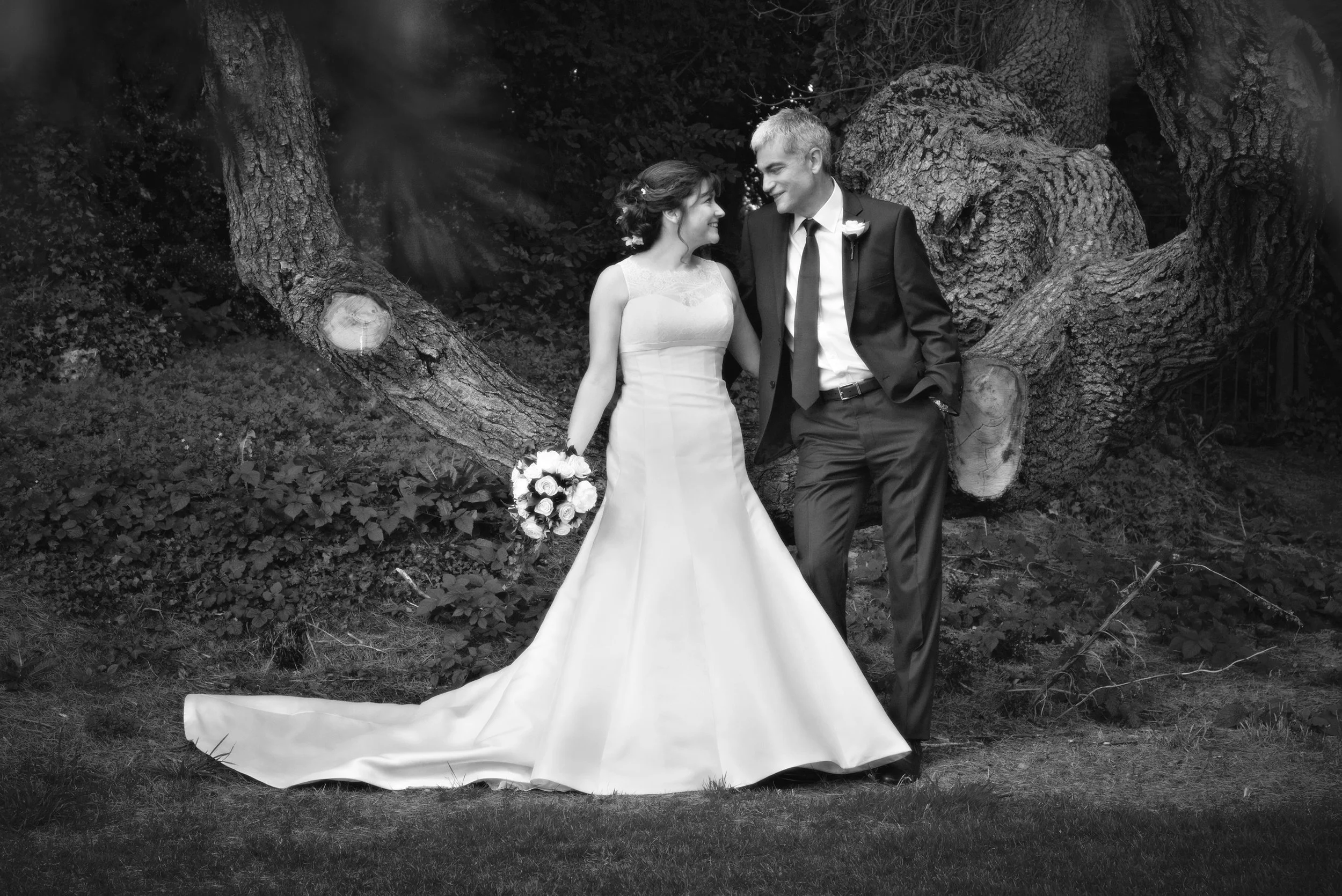 A black and white photograph of a bride and groom standing outdoors next to a large tree with textured bark. The bride is wearing a sleeveless wedding gown and holding a bouquet of roses, while the groom is dressed in a dark suit and tie. They are ga