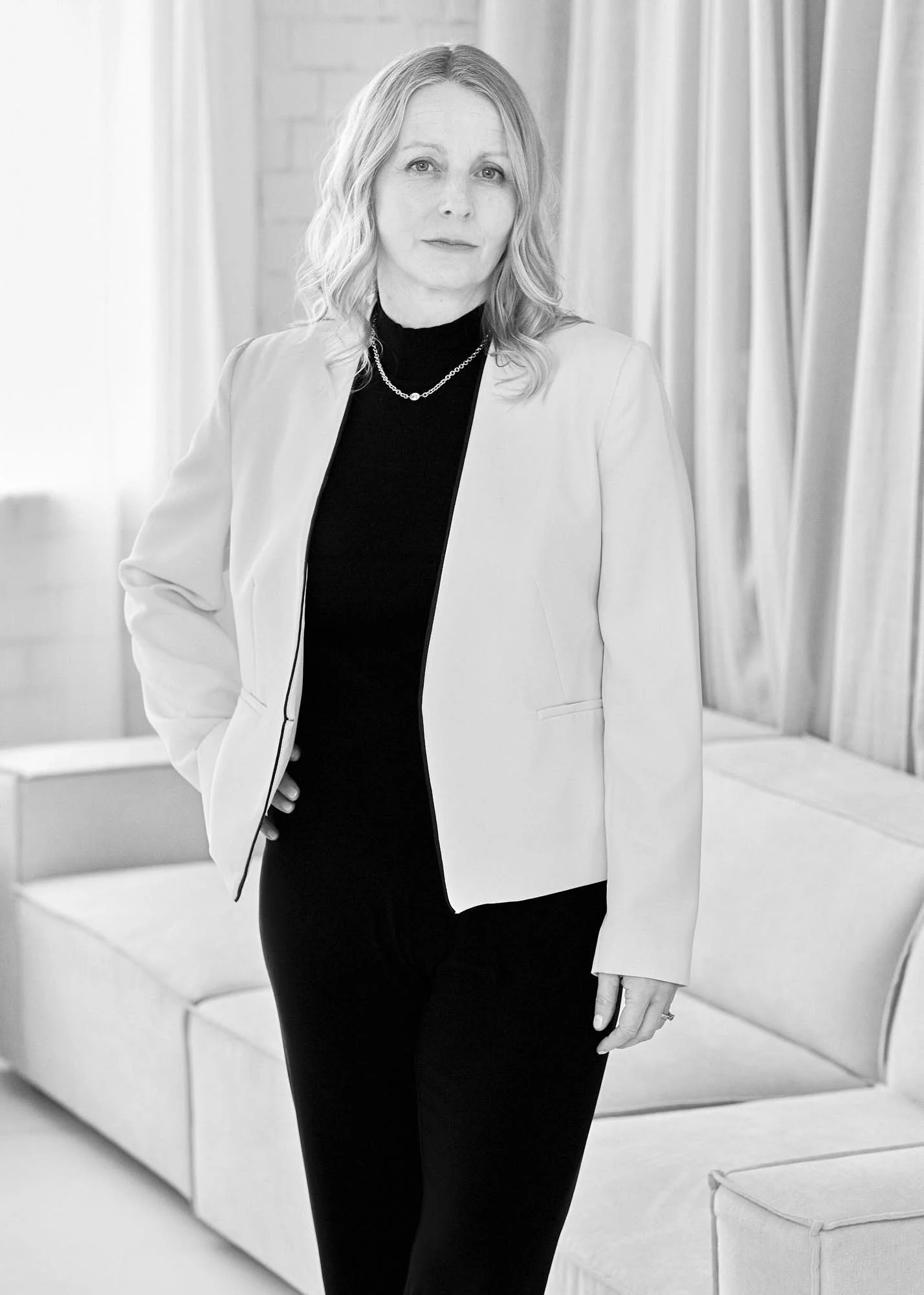 Black and white photo of a woman with shoulder-length wavy hair, wearing a light-colored blazer, black top, and layered necklaces, standing in a modern room with curtains and seating furniture.