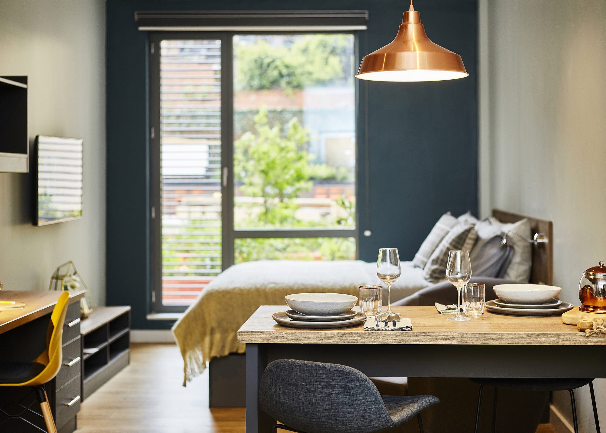 A modern dining room with a wooden table set with bowls, plates, napkins, and glasses. There are dark chairs around the table, and a blue accent wall in the background with a large window showing greenery outside. A copper pendant light hangs above the table.