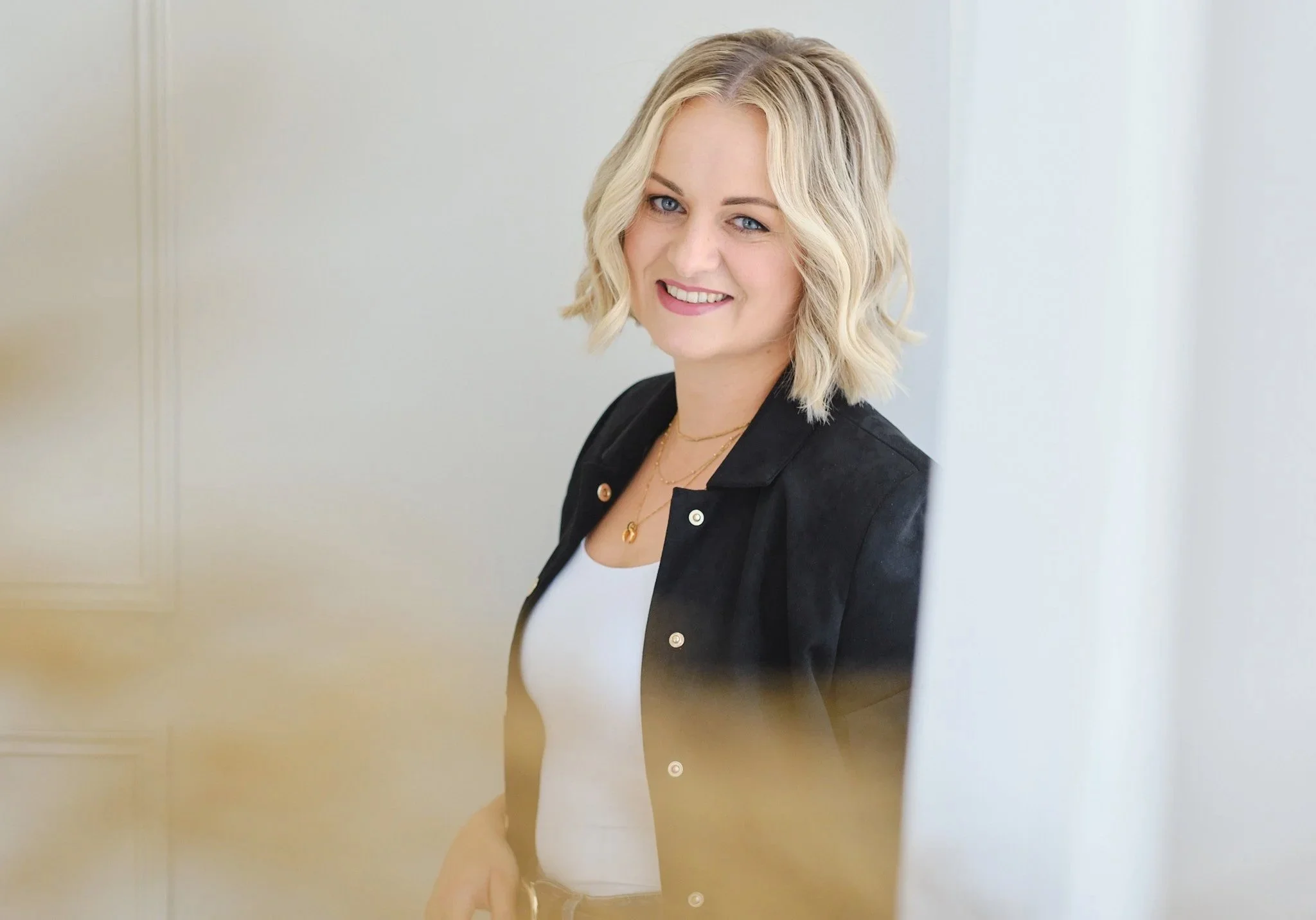 A smiling woman with blonde hair and blue eyes, wearing a black jacket and gold jewelry, standing near a white wall.