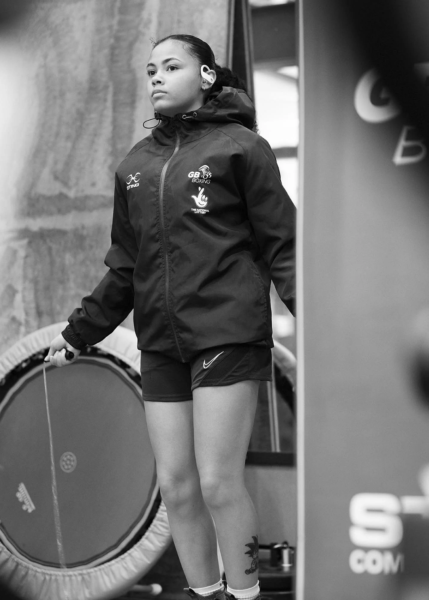 A young girl in athletic wear, including a jacket with logos and shorts, holding a jump rope, standing indoors near exercise equipment, with a serious expression.