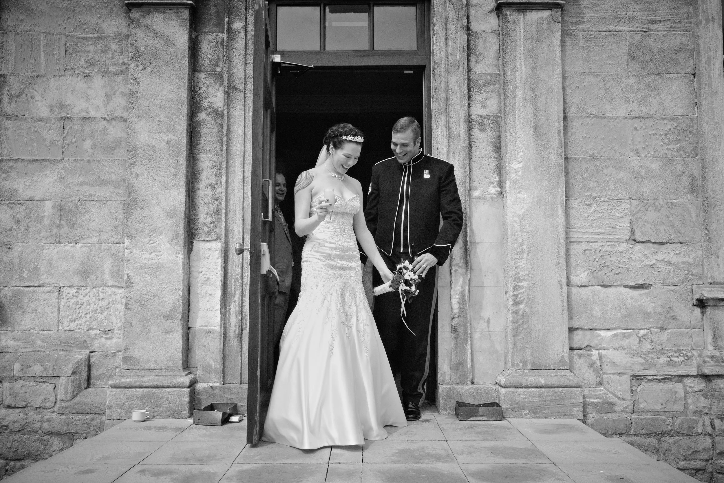 A bride and groom emerging from a stone church doorway, smiling and looking at each other. The bride is in a strapless wedding gown and holding a small bouquet, while the groom is in a formal uniform. An officiant and a guest are visible in the backg