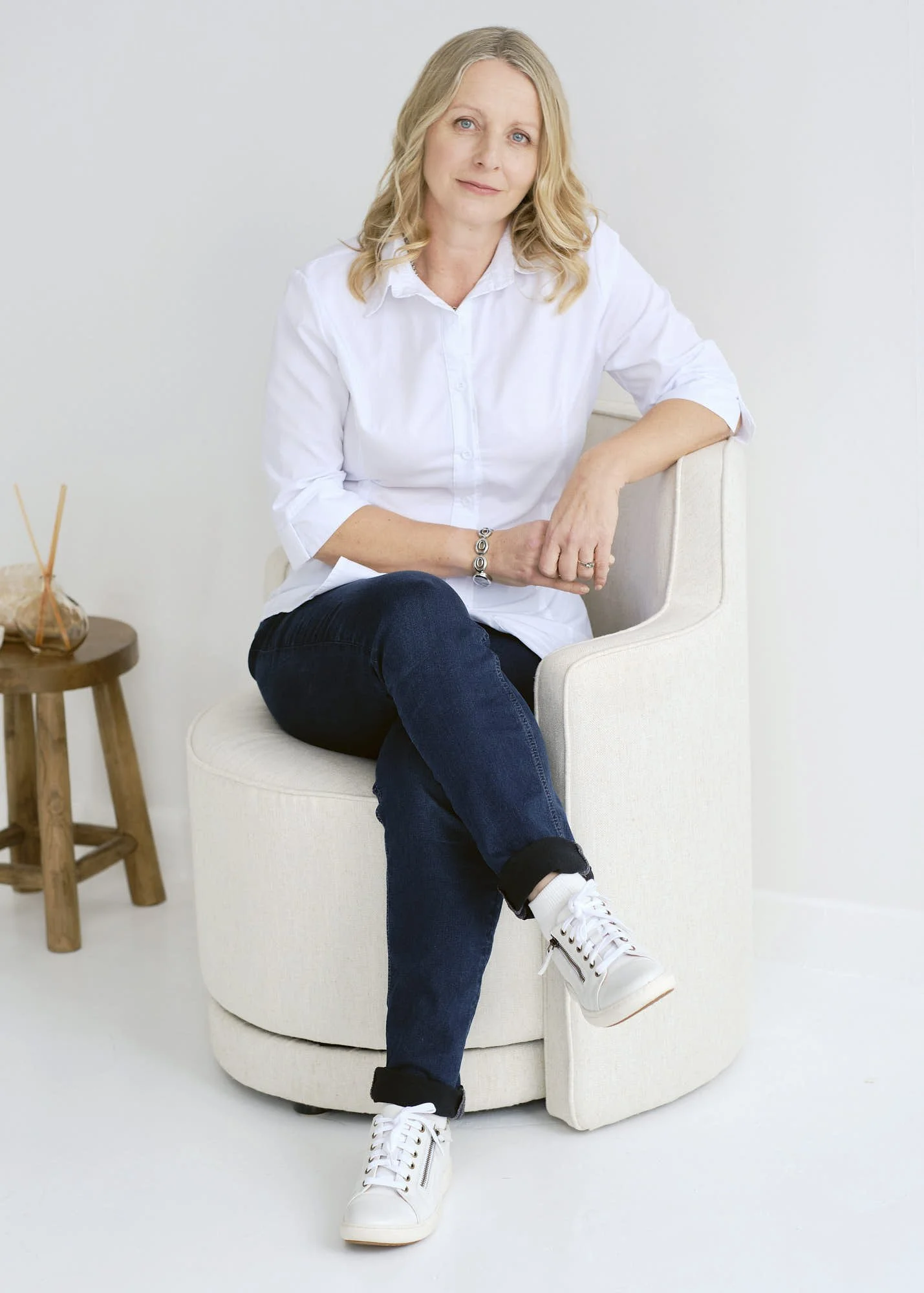 A woman with blonde hair wearing a white shirt, dark jeans, and white sneakers sitting on a beige chair with one leg crossed over the other. In the background, there is a small wooden side table with a decorative object.