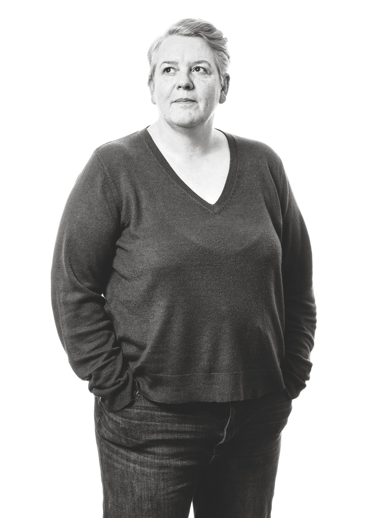 Black and white photo of a woman with short hair wearing a V-neck sweater and dark jeans, standing with one hand in her pocket against a plain white background.