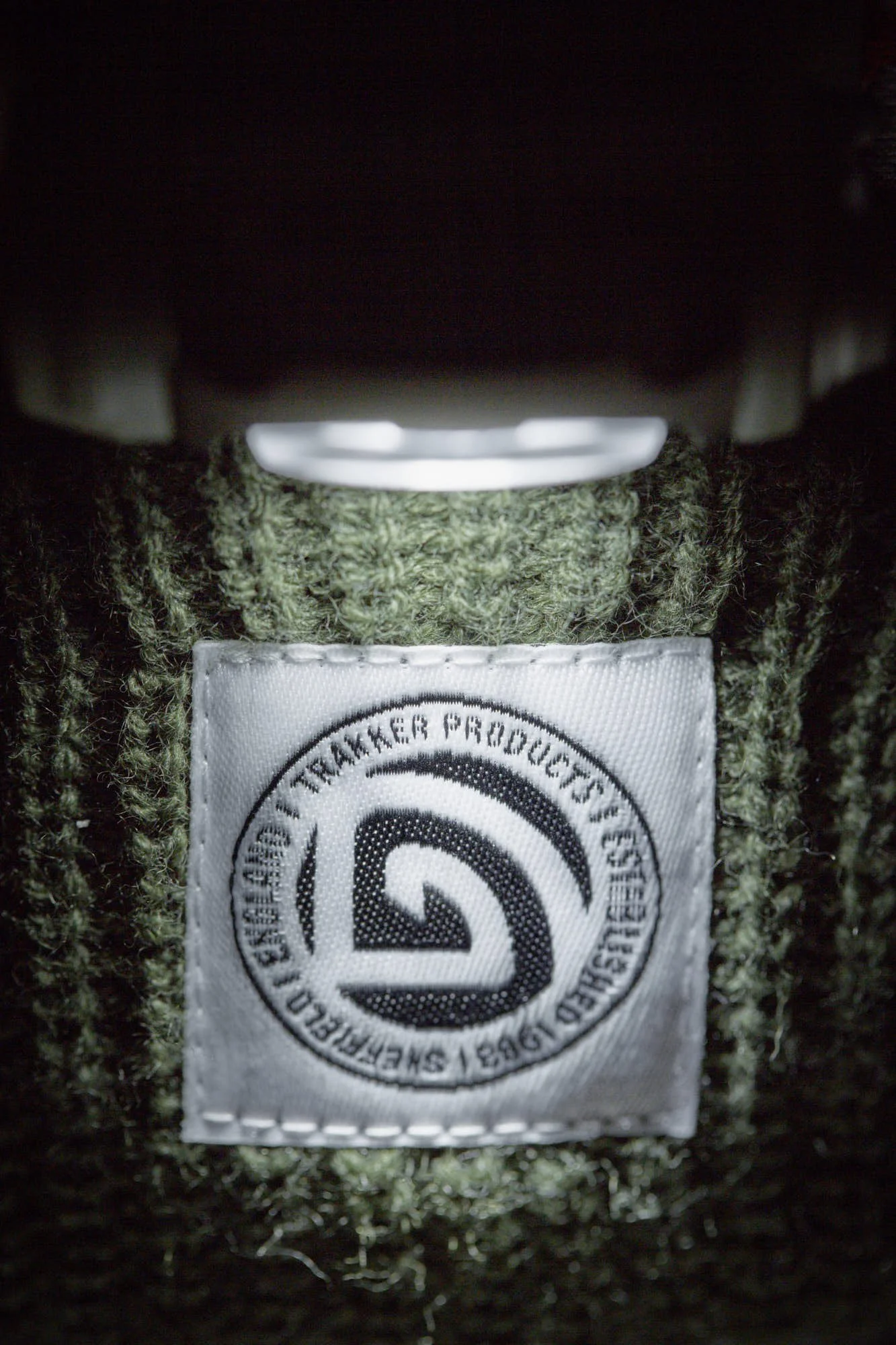 Close-up of a white fabric tag with a circular logo and text, attached to a green, fuzzy fabric material, underneath a dark surface.