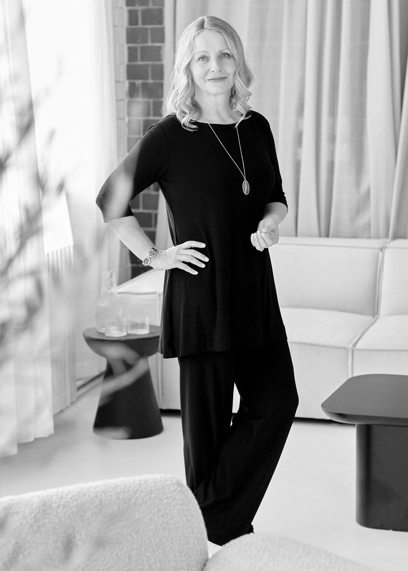 A woman with shoulder-length wavy hair wearing a black dress with a pendant necklace and bracelet, standing in a room with curtains, a brick wall, and modern furniture.