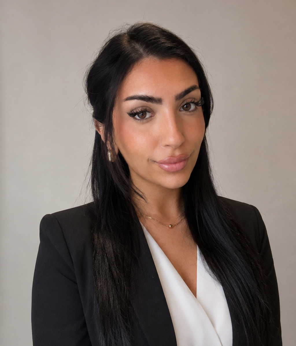A professional woman with long dark hair, wearing a black blazer and white blouse, posing against a plain light-colored background.