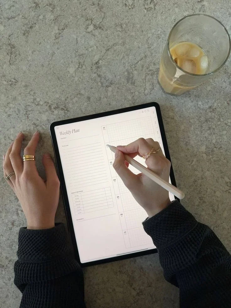 Person writing in a weekly planner on a tablet with a stylus, with a glass of iced beverage nearby on a textured surface.