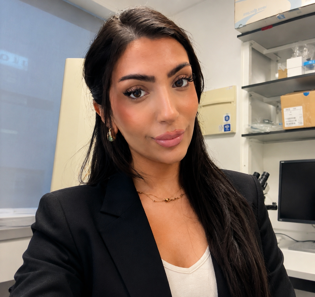 A young woman with long dark hair, wearing makeup, gold jewelry, a black blazer, and a white top, sitting in a laboratory or office setting.