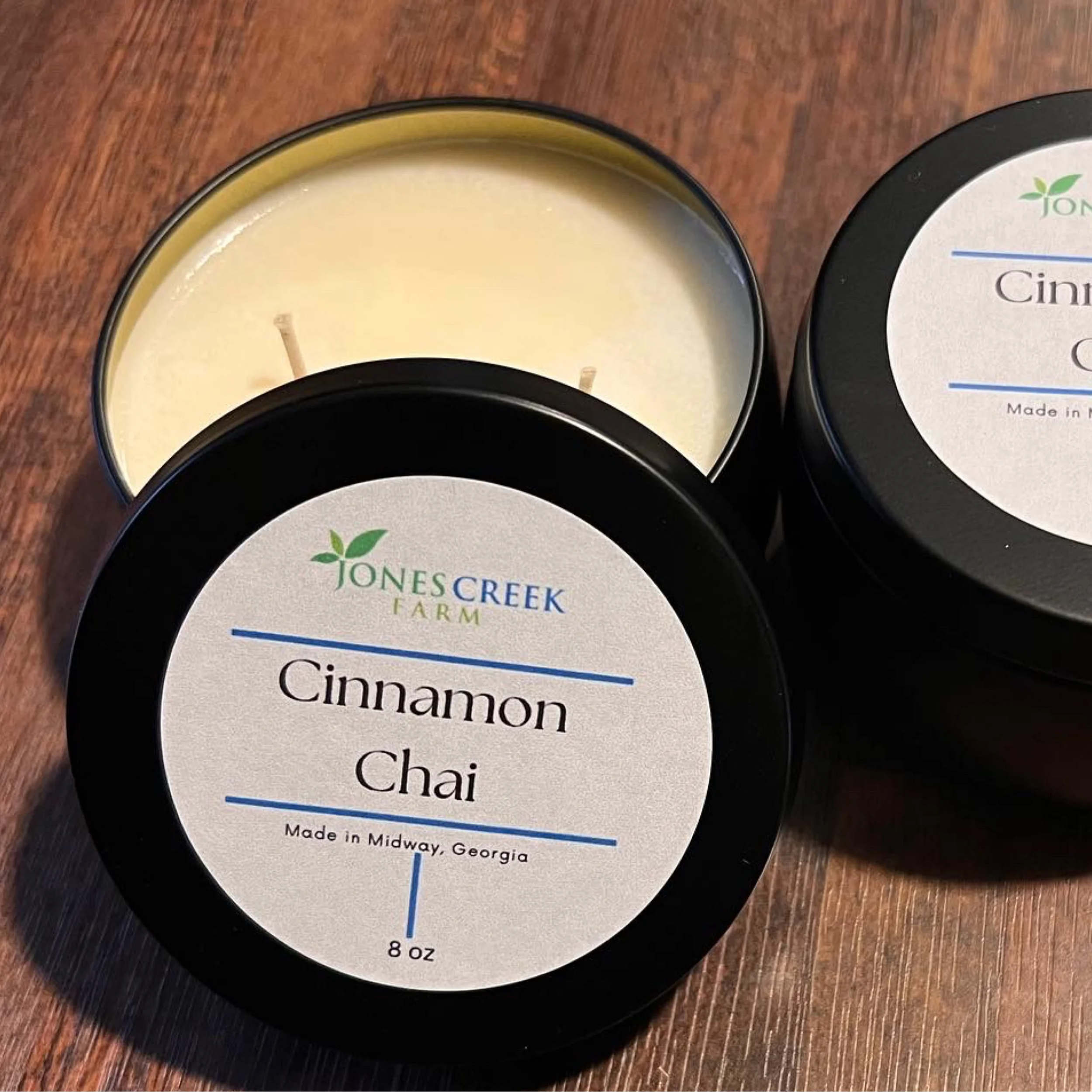 A close-up of several scented candles in round tins with black lids, on a wooden surface. The label on the candle reads "Cinnamon Chai" and indicates it is 8 ounces and made in Midway, Georgia.