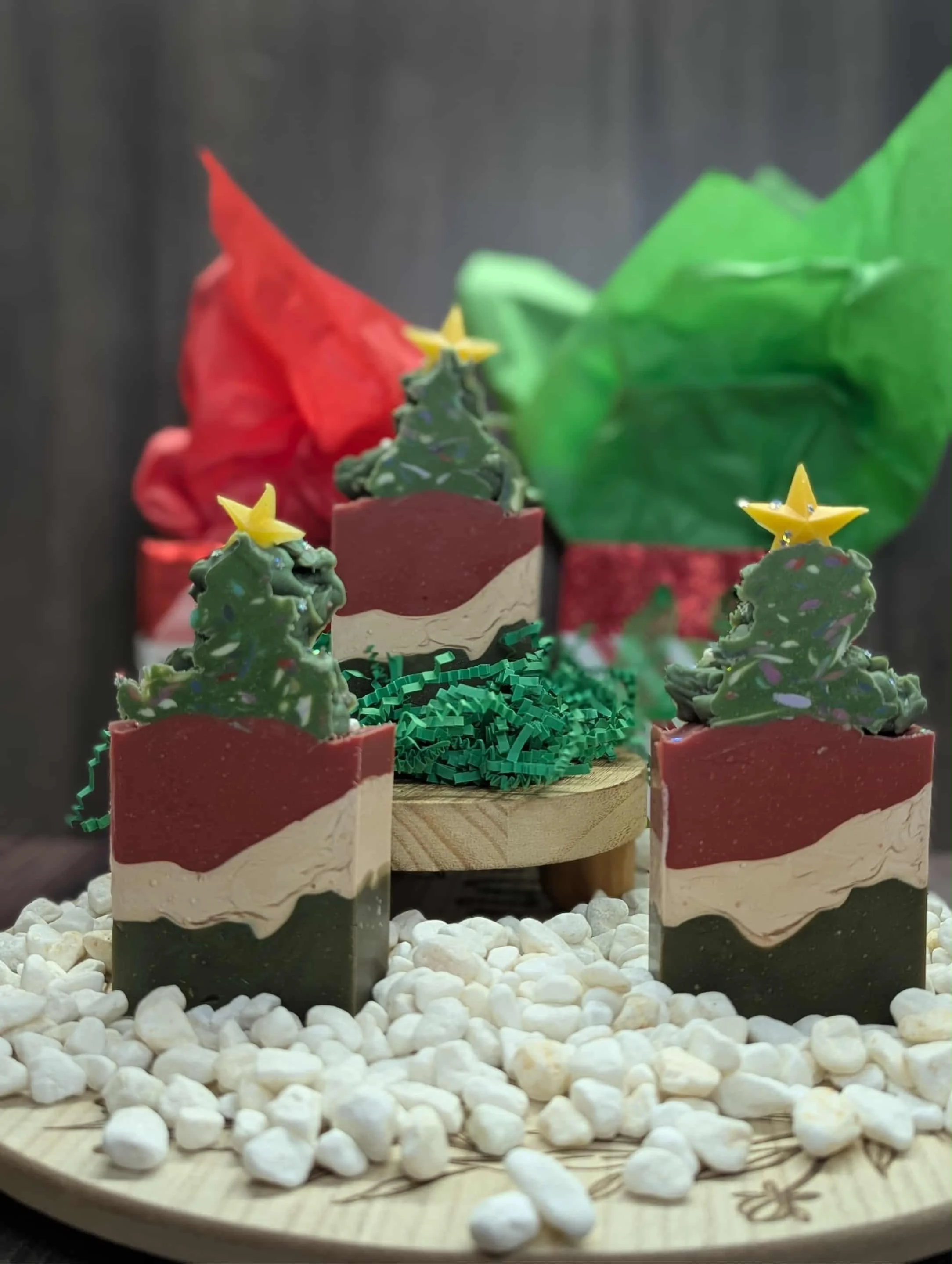 Small decorative Christmas trees on a wooden tray, with a red, green, and cream-colored cake or decoration behind them, surrounded by white pebbles.