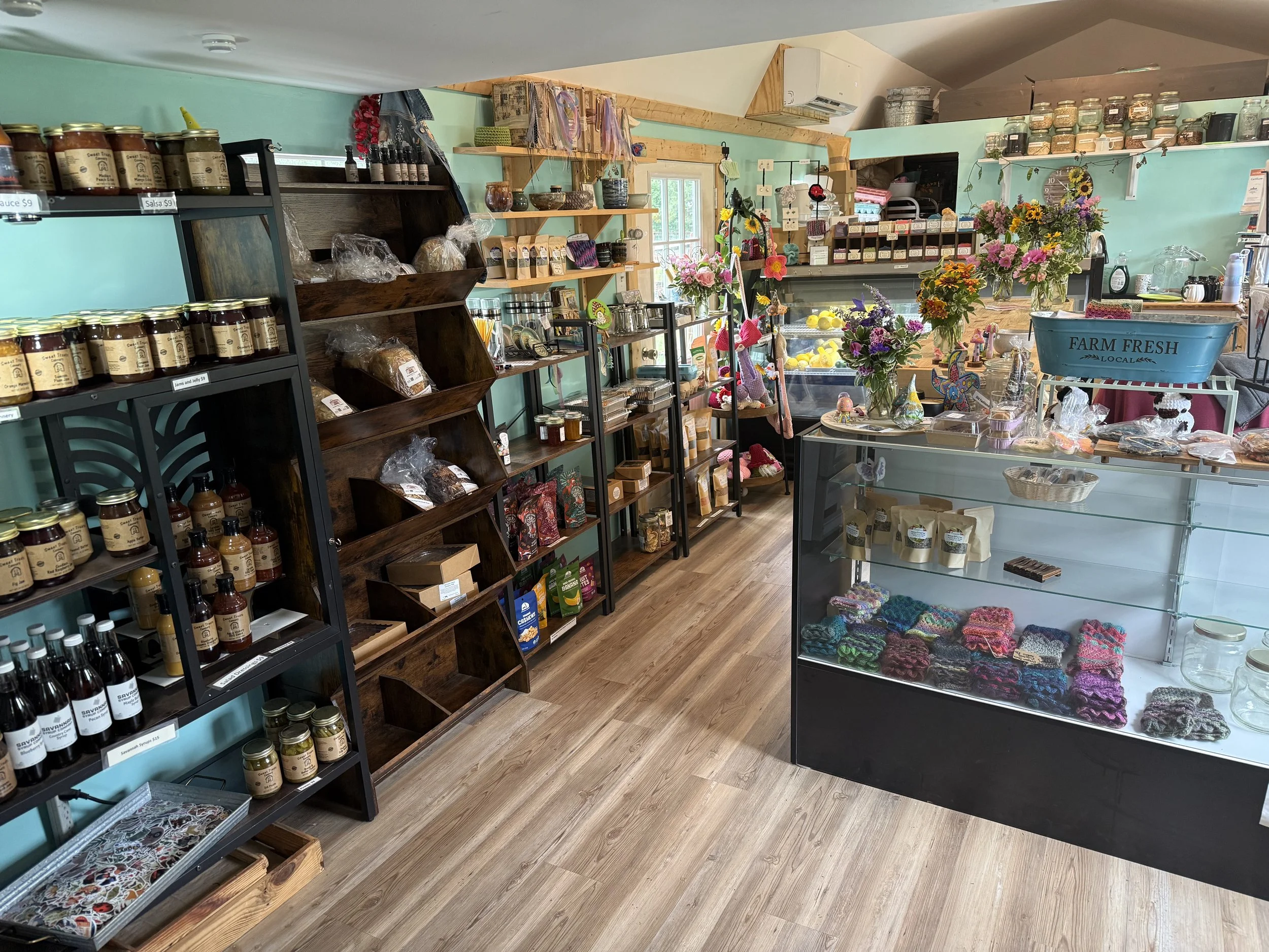 Interior view of a small shop with shelves and display cases filled with jars, bottles, handmade crafts, and flowers, located under a sloped ceiling with wooden accents and a window in the background.