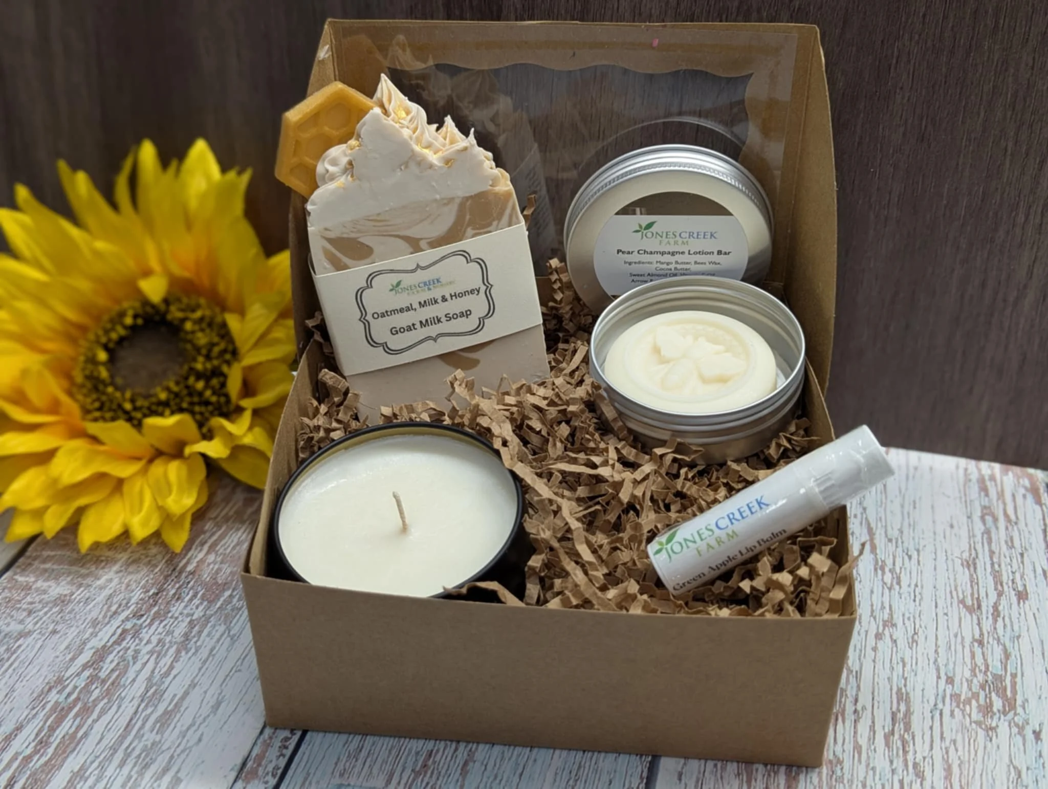 A gift box containing natural skincare products from Jones Creek Farm, including a soap labeled 'Oatmeal, Milk & Honey Goat Milk Soap' and two tin containers of lotions, with a yellow sunflower on the left side of the image.